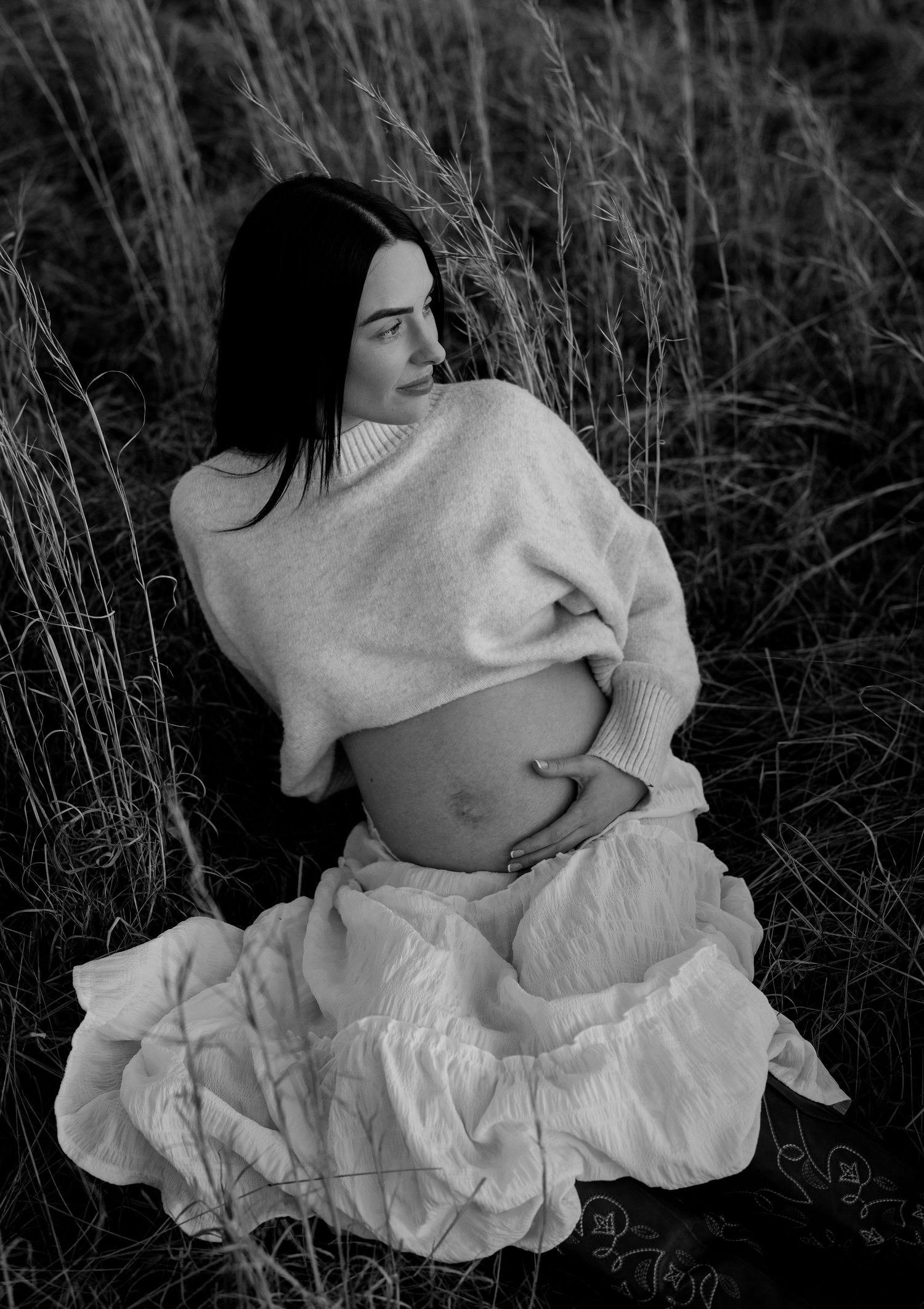 Black and white maternity photo of pregnant woman sitting in tall grass holding her belly.