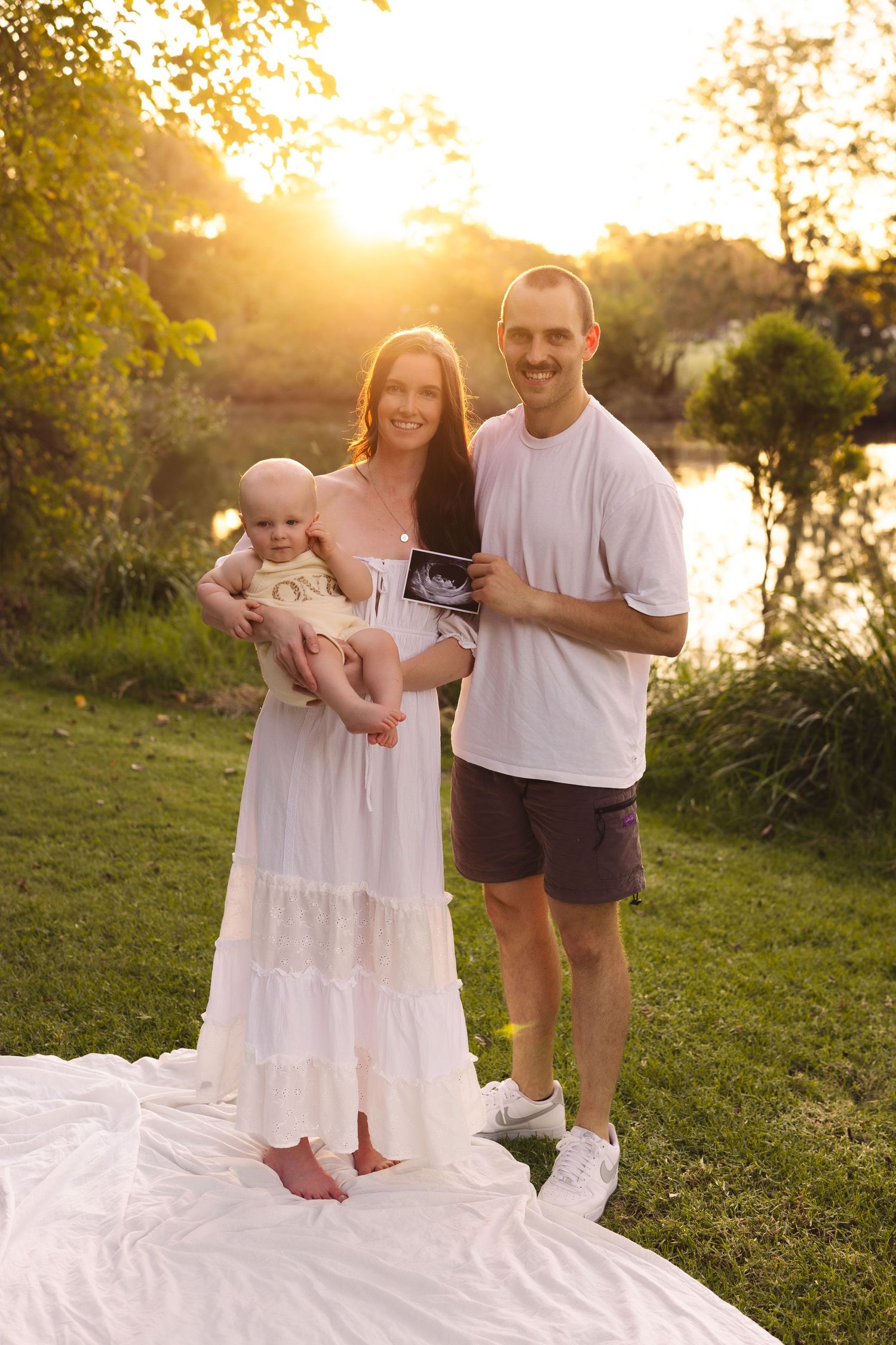 Happy family posing outdoors at golden hour, mother holding baby, father displaying ultrasound photo near a serene pond.