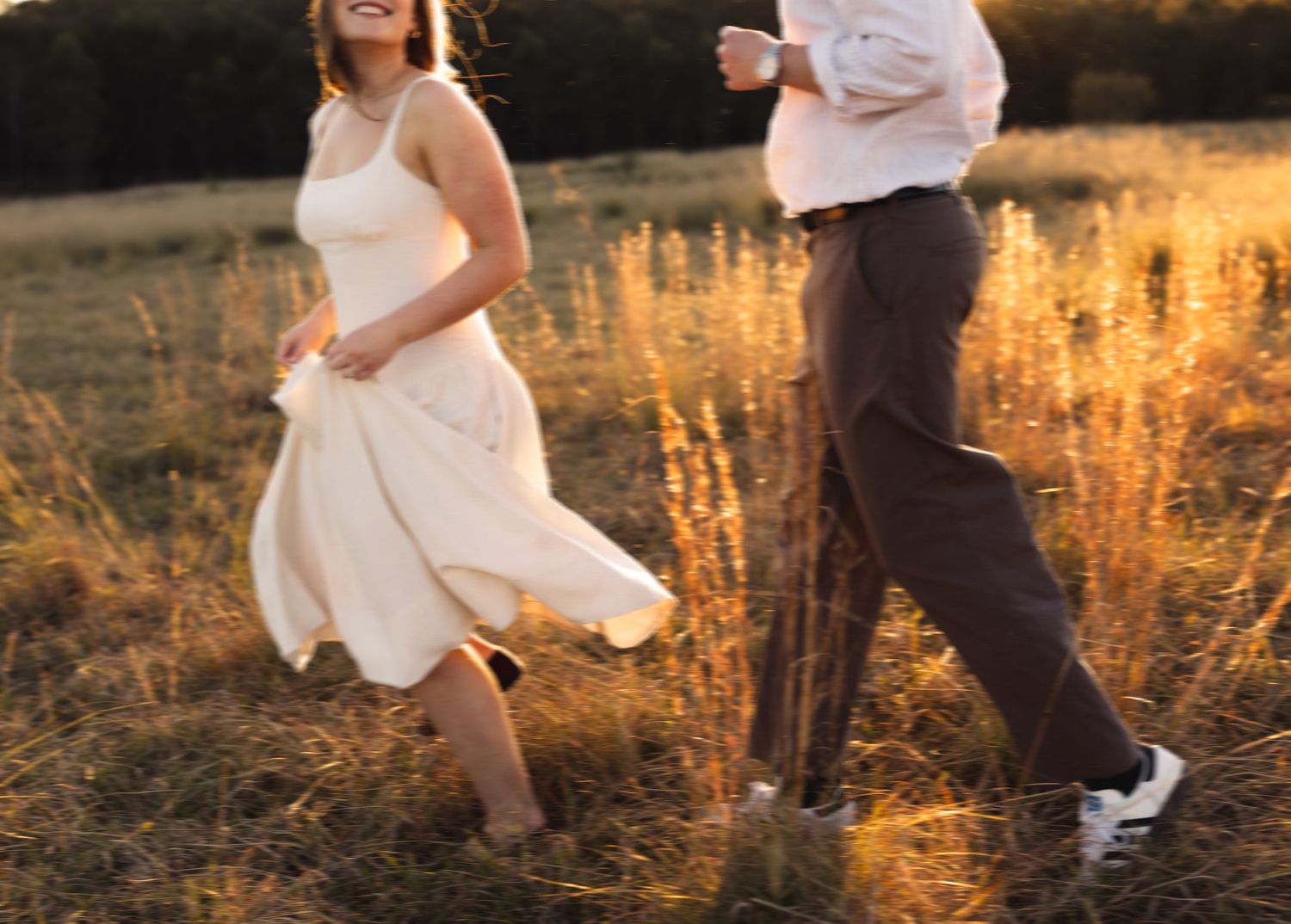 Joyful couple running through golden sunlit field at sunset, woman in flowing white dress laughing freely.
