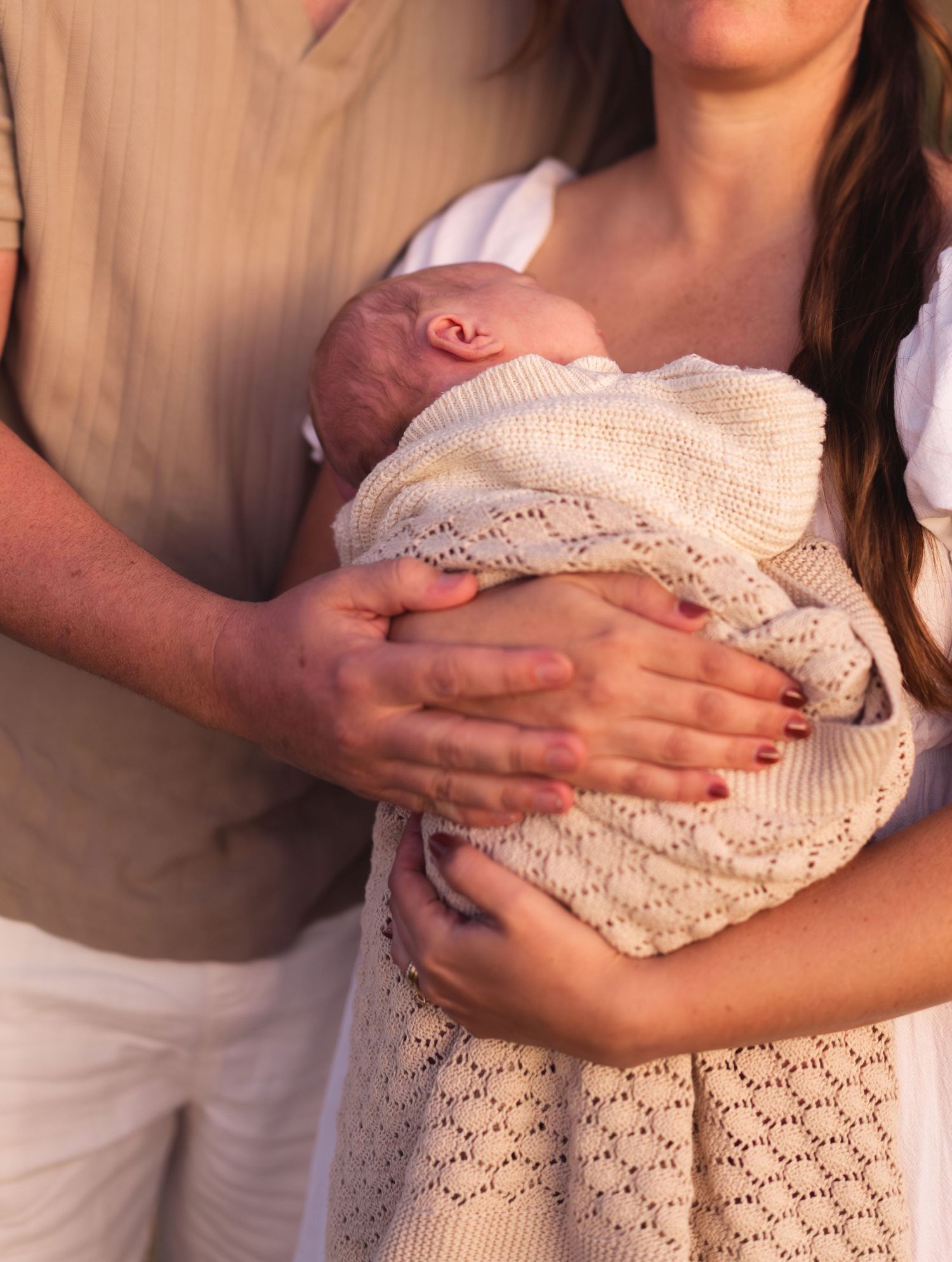 Parents cradling newborn baby wrapped in white knit blanket outdoors.
