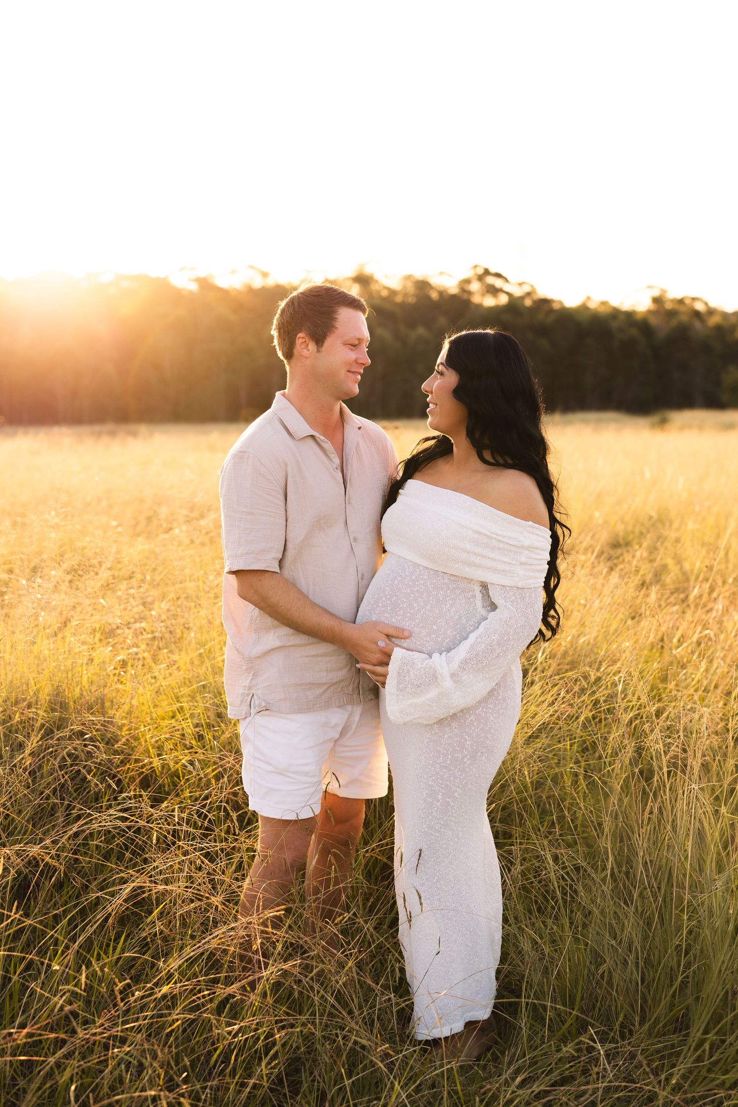 Couple sharing a tender moment in golden field at sunset, pregnant woman in white lace gown, man in linen shirt.