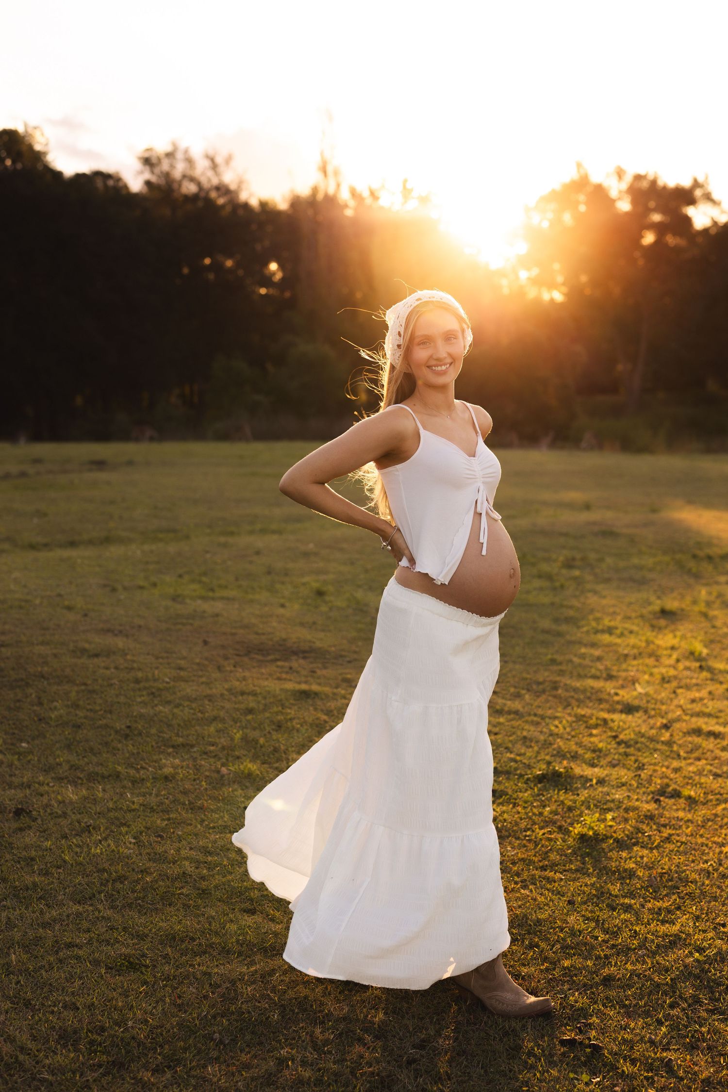 Pregnant woman in white dress glowing at golden sunset in open field.