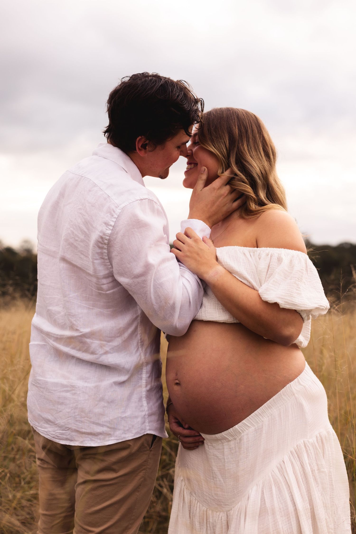 Expecting couple sharing intimate moment in golden field, woman in white off-shoulder dress.