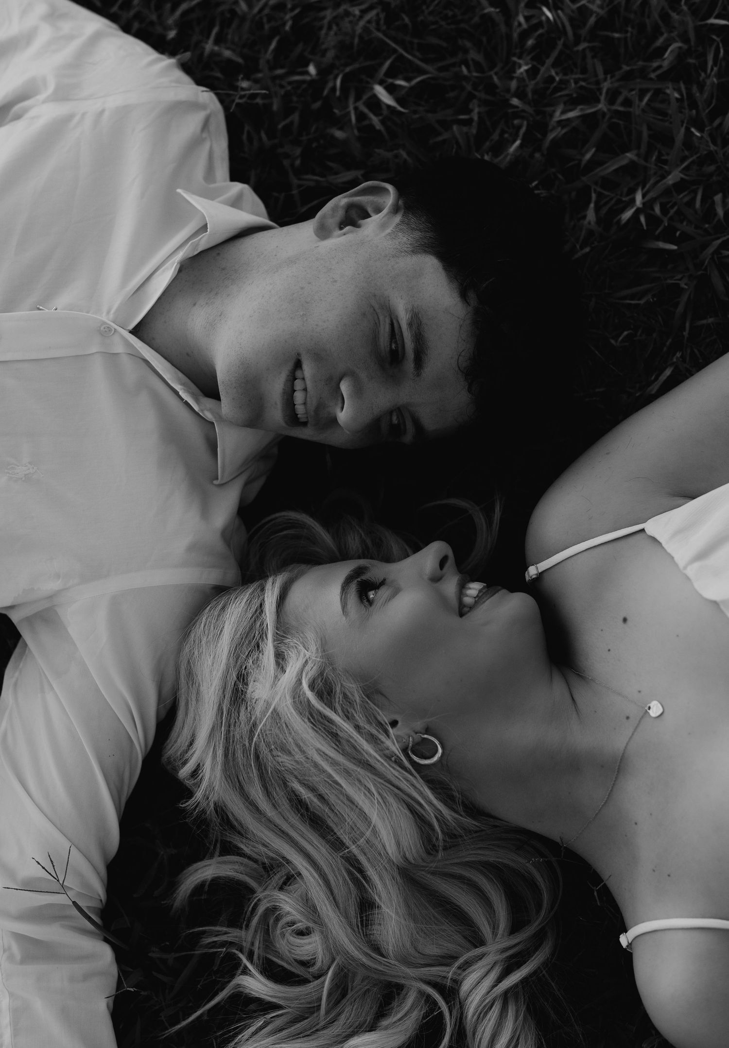 Black and white photo of a smiling couple lying on grass looking at each other romantically.