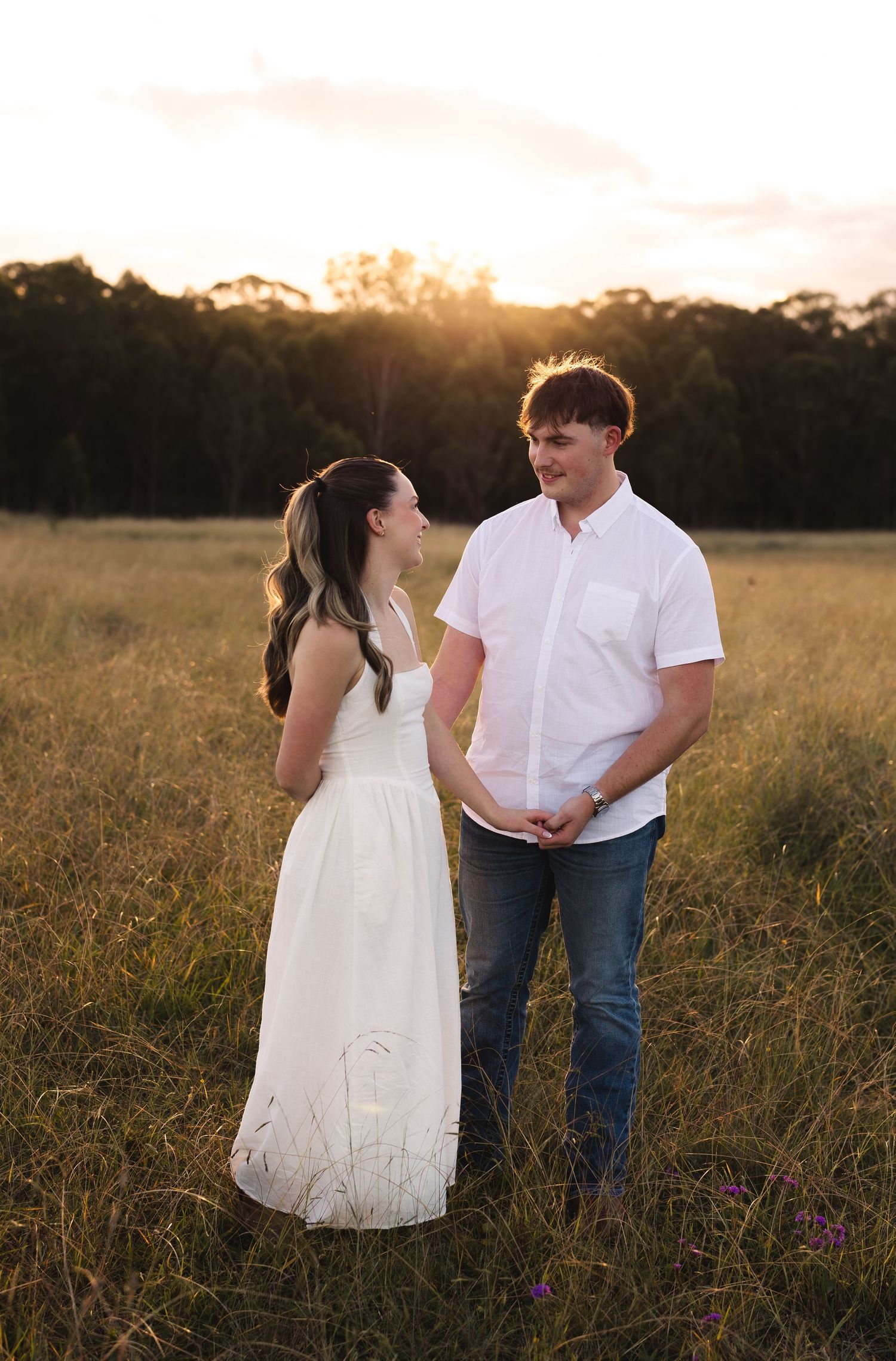 Young couple holding hands and smiling at each other in a golden field during sunset.