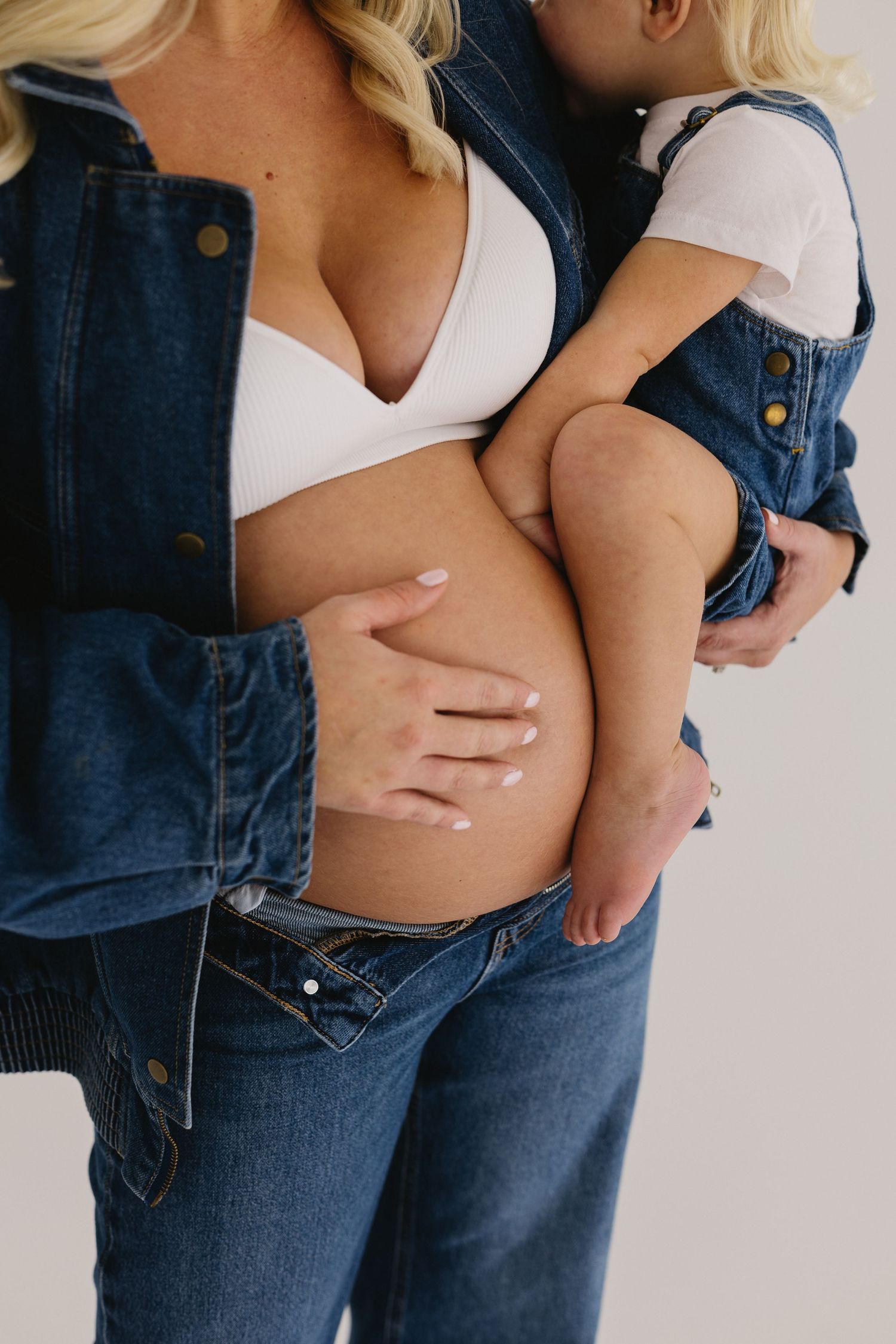 Pregnant woman in denim holding toddler child against bare baby bump.