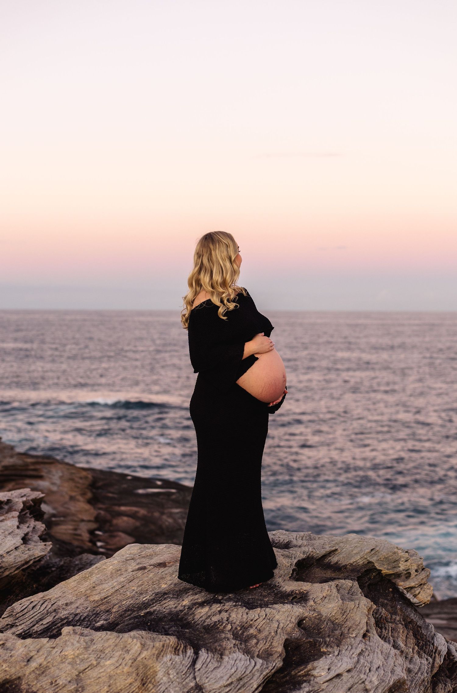 Pregnant woman in black gown cradling baby bump on rocky shore at sunset.