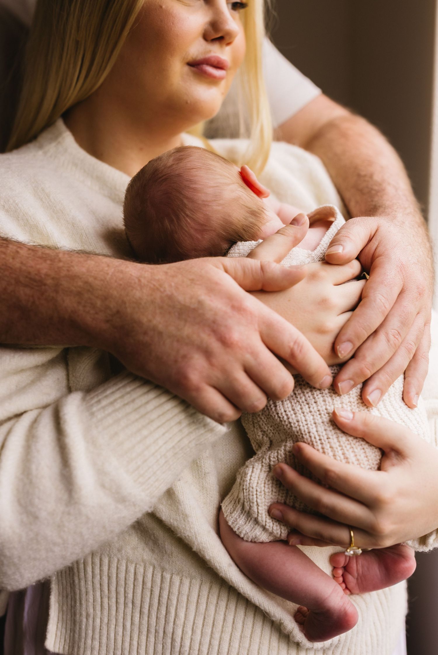 Parents tenderly holding newborn baby together, mother in cream sweater, father with wedding ring.