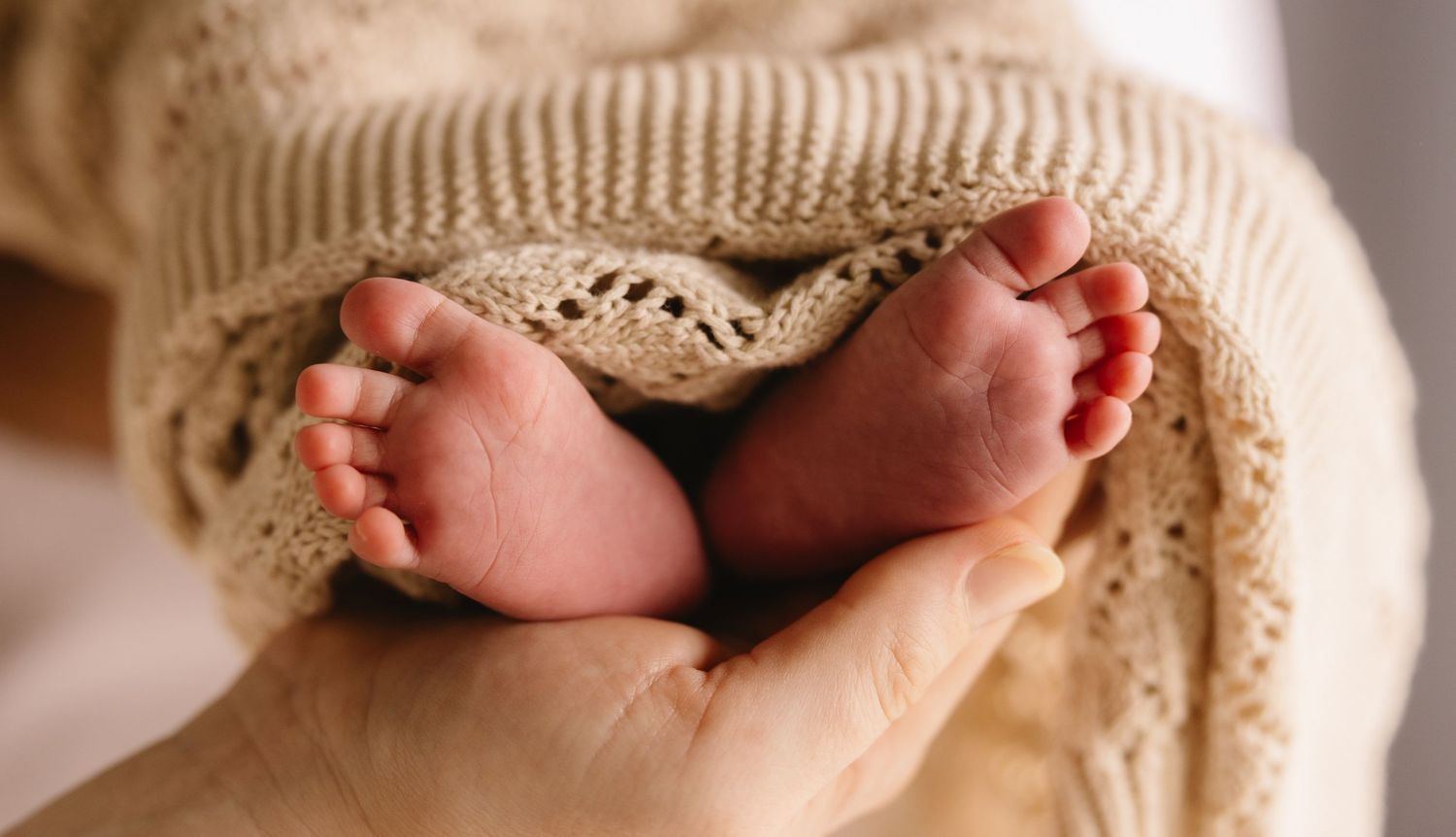 Tiny newborn baby feet cradled gently in adult hands with knit blanket.