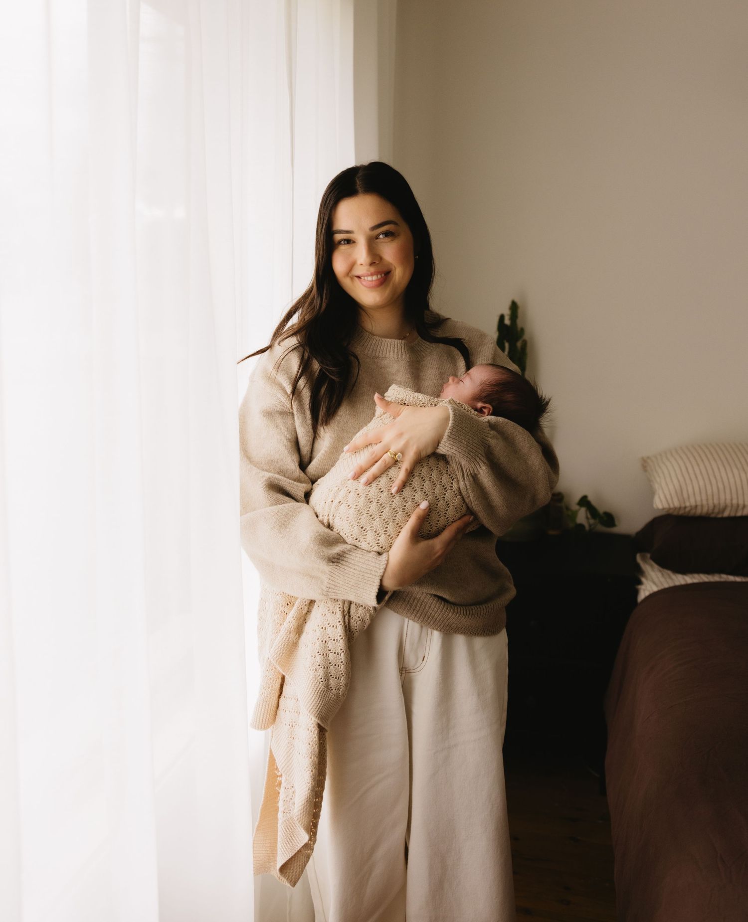 Woman in beige sweater holding sleeping newborn wrapped in knit blanket near bright window.