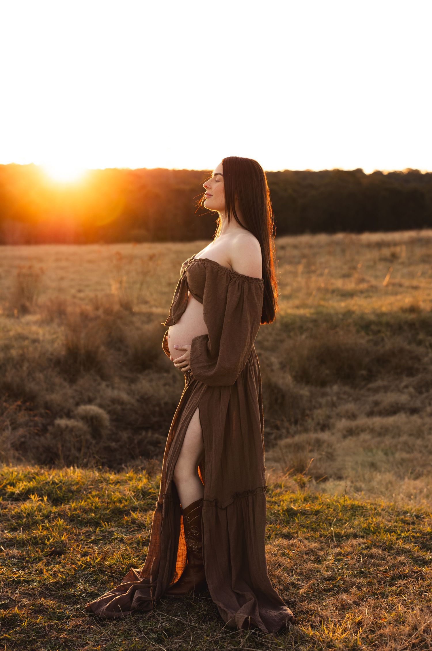 Pregnant woman in brown off-shoulder dress holding belly at golden sunset in open field.