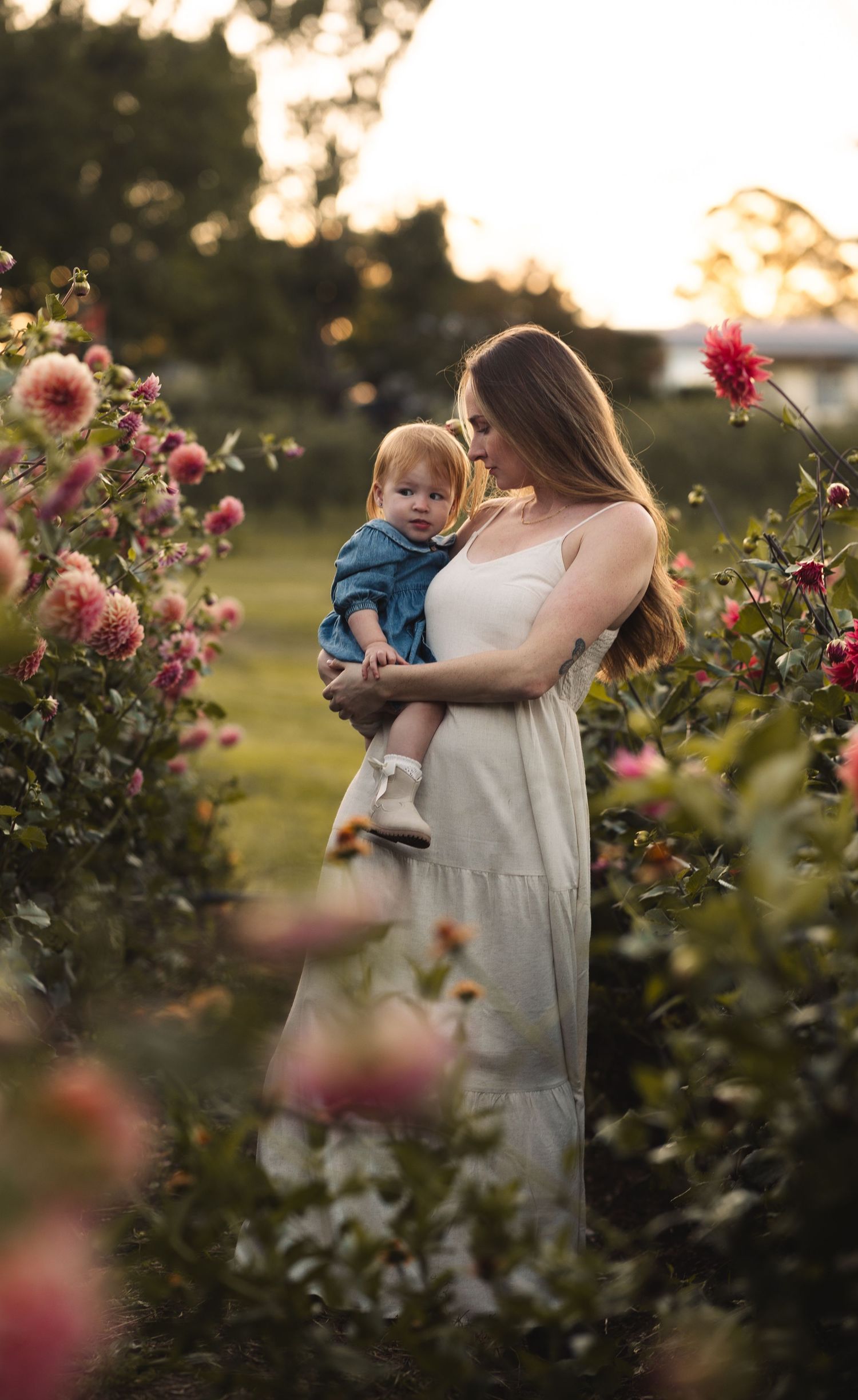 Mother in white dress holding baby among blooming pink flowers at golden hour.