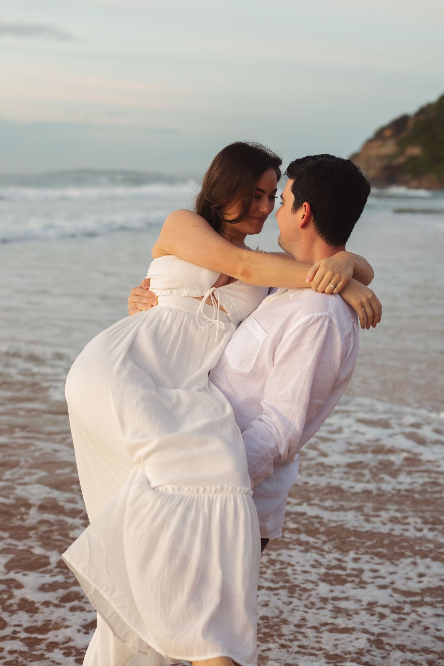 Romantic couple in white outfits embracing on a sandy beach at sunset with ocean waves in the background.