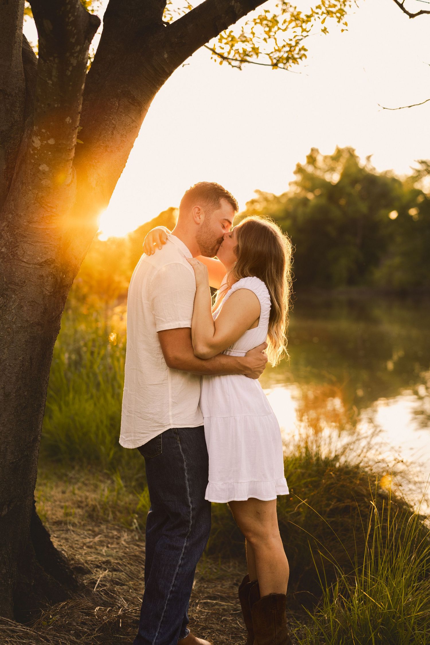 Couple kissing at golden sunset by a lake under a tree, woman in white dress and boots.