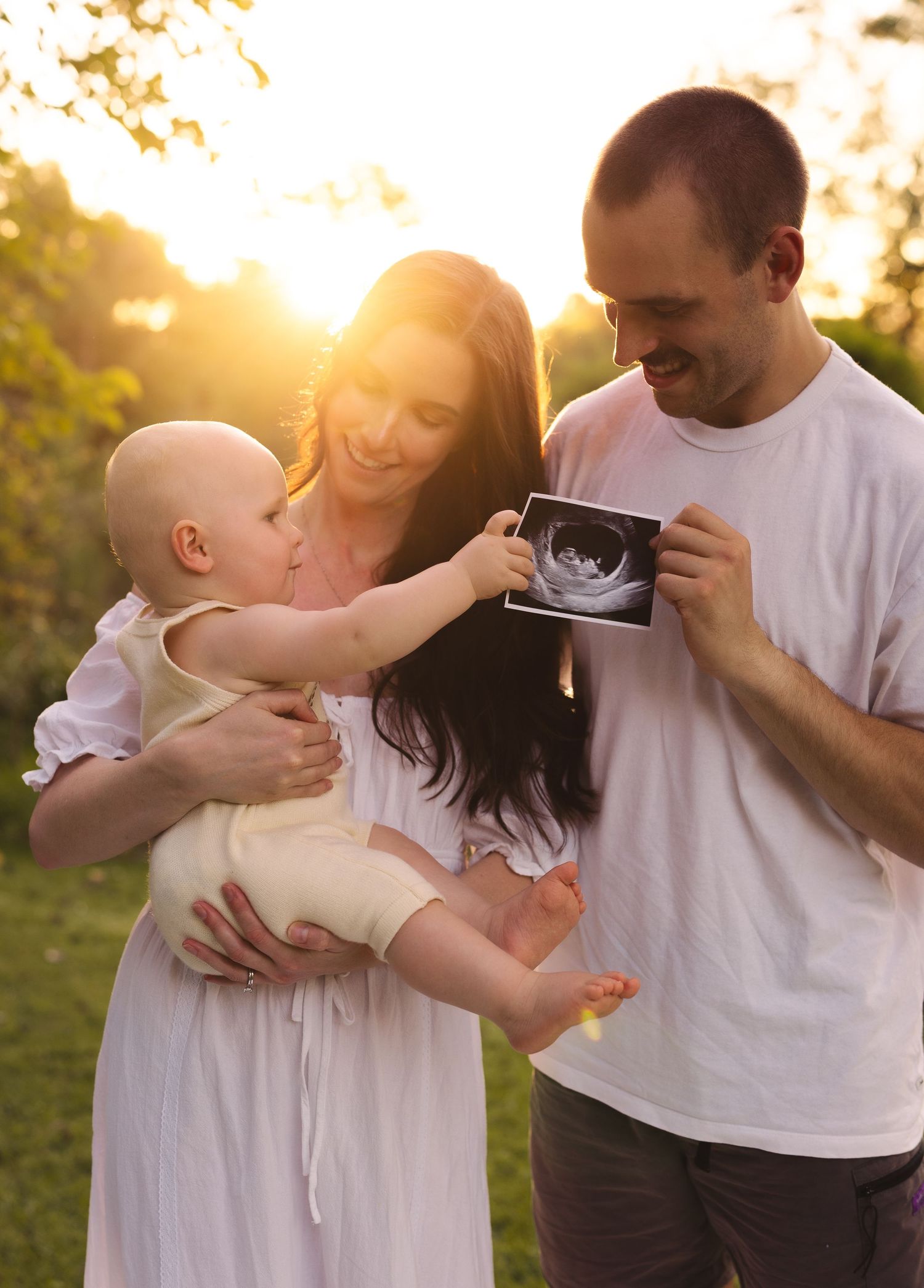 Happy couple holding baby and ultrasound photo in sunny garden during pregnancy announcement.