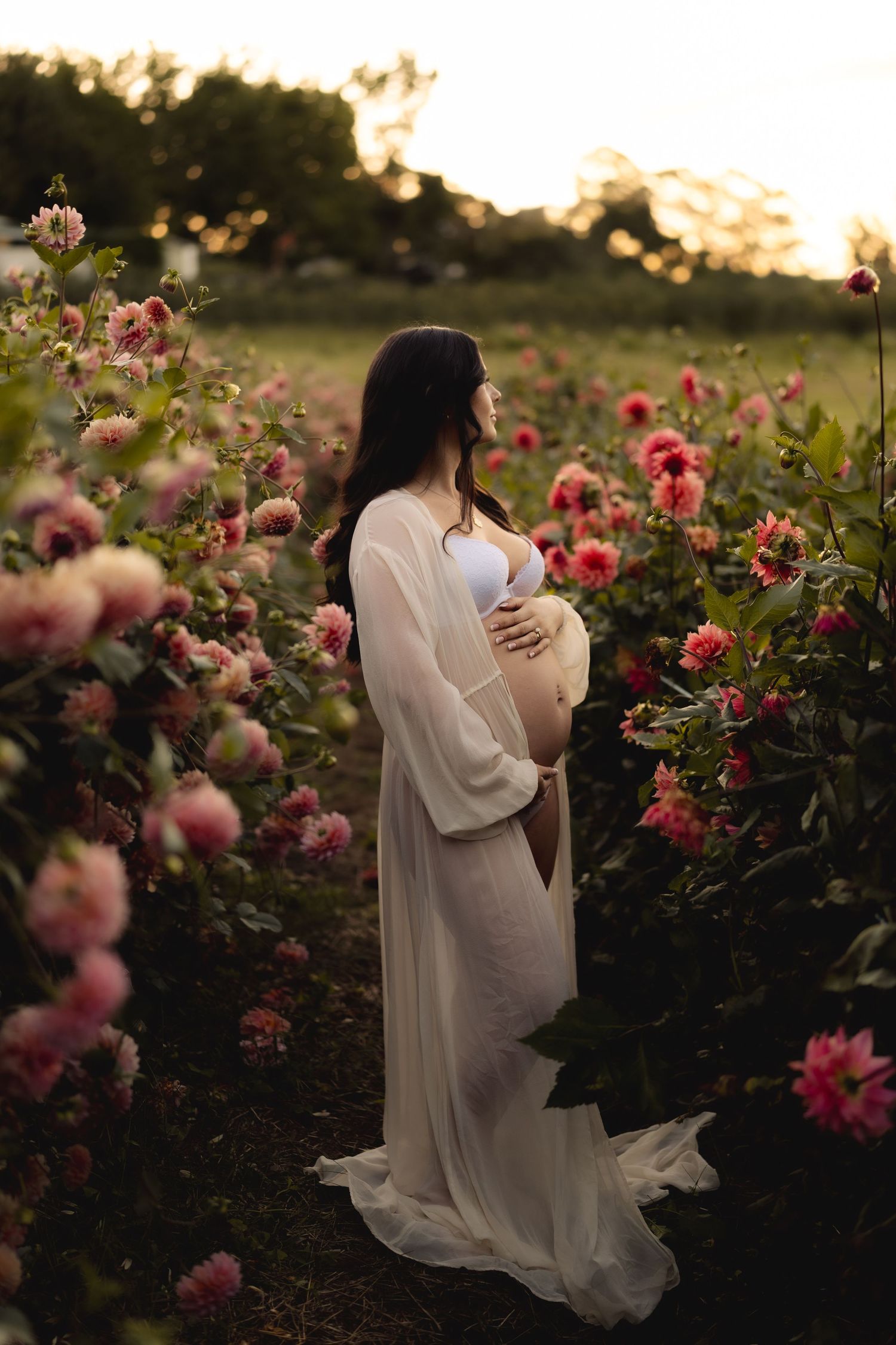 Pregnant woman in white sheer gown standing among vibrant pink dahlias at dusk.
