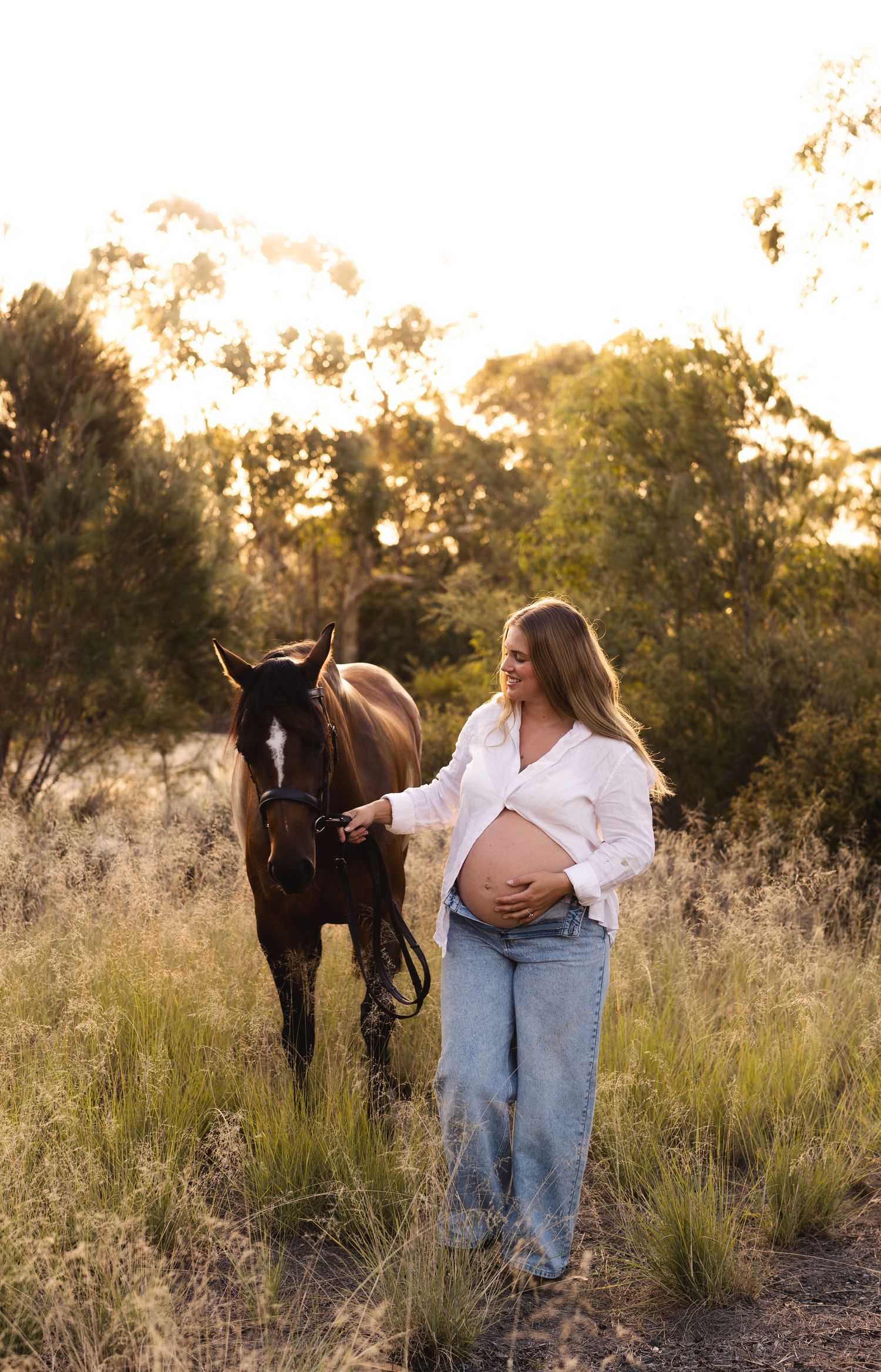 Smiling pregnant woman in white shirt and jeans walking with a horse at golden sunset.