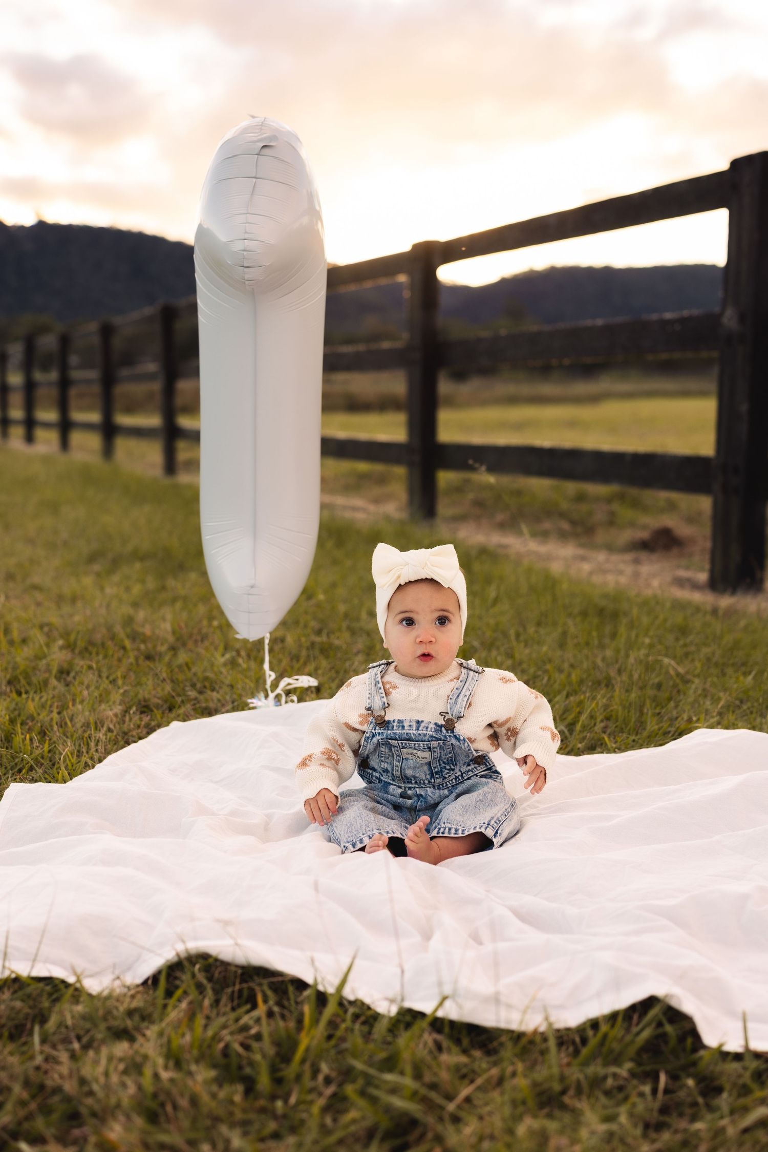 Baby in denim overalls sitting on white blanket with balloon at sunset.