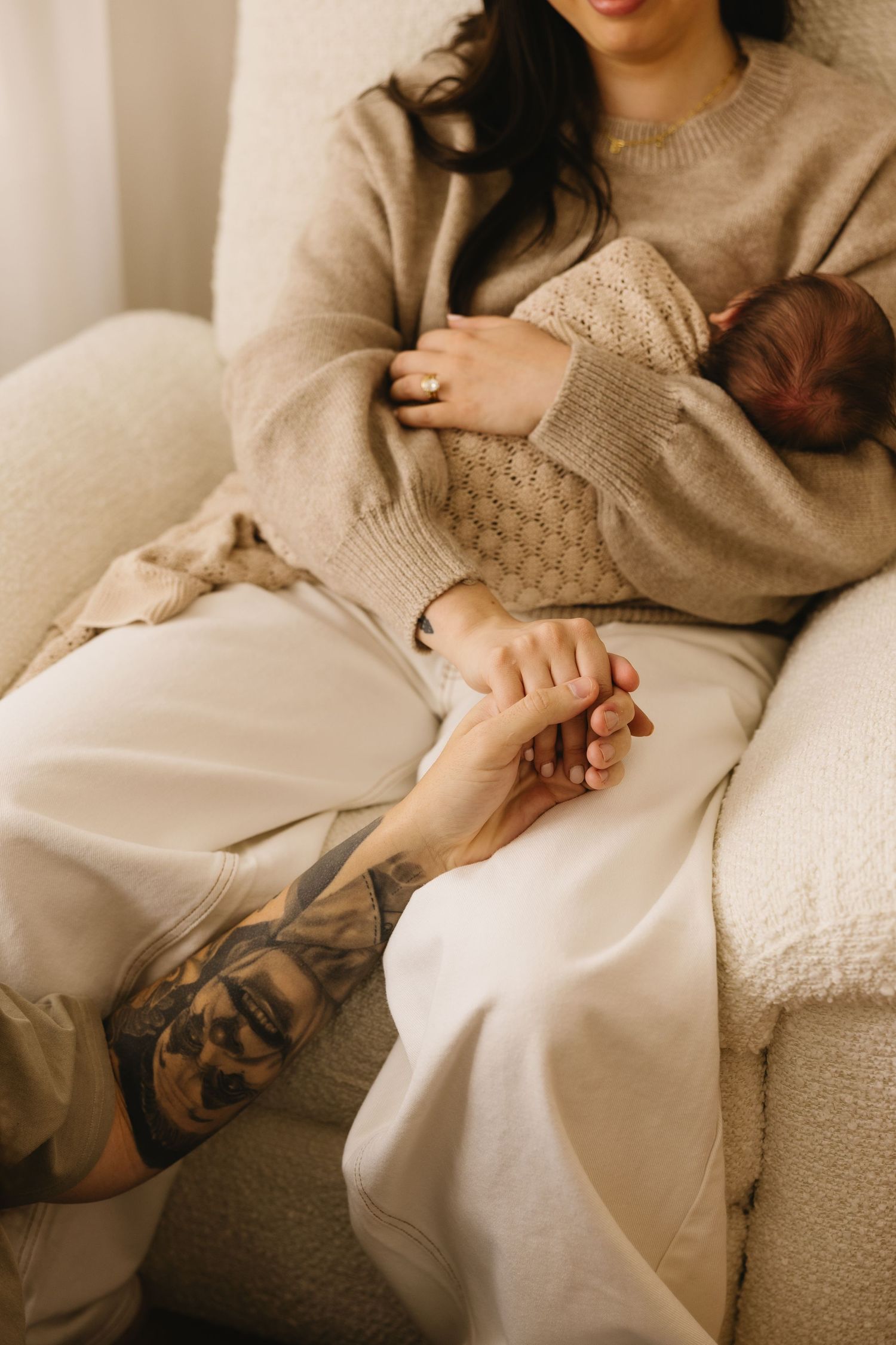 Mother in beige sweater holds newborn while partner with tattoos holds her hand.