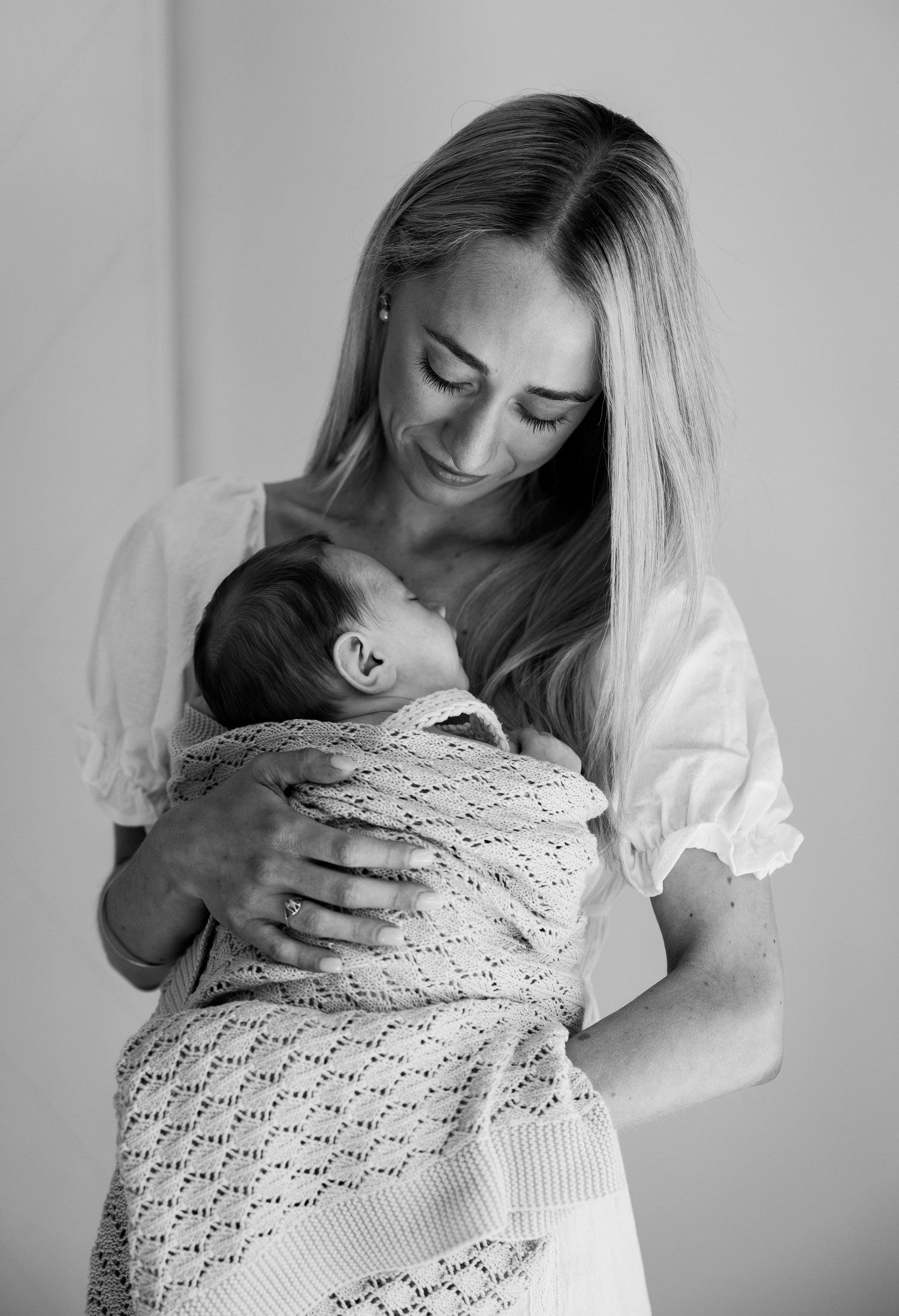 Smiling blonde mother in white dress cradles swaddled newborn in black and white.