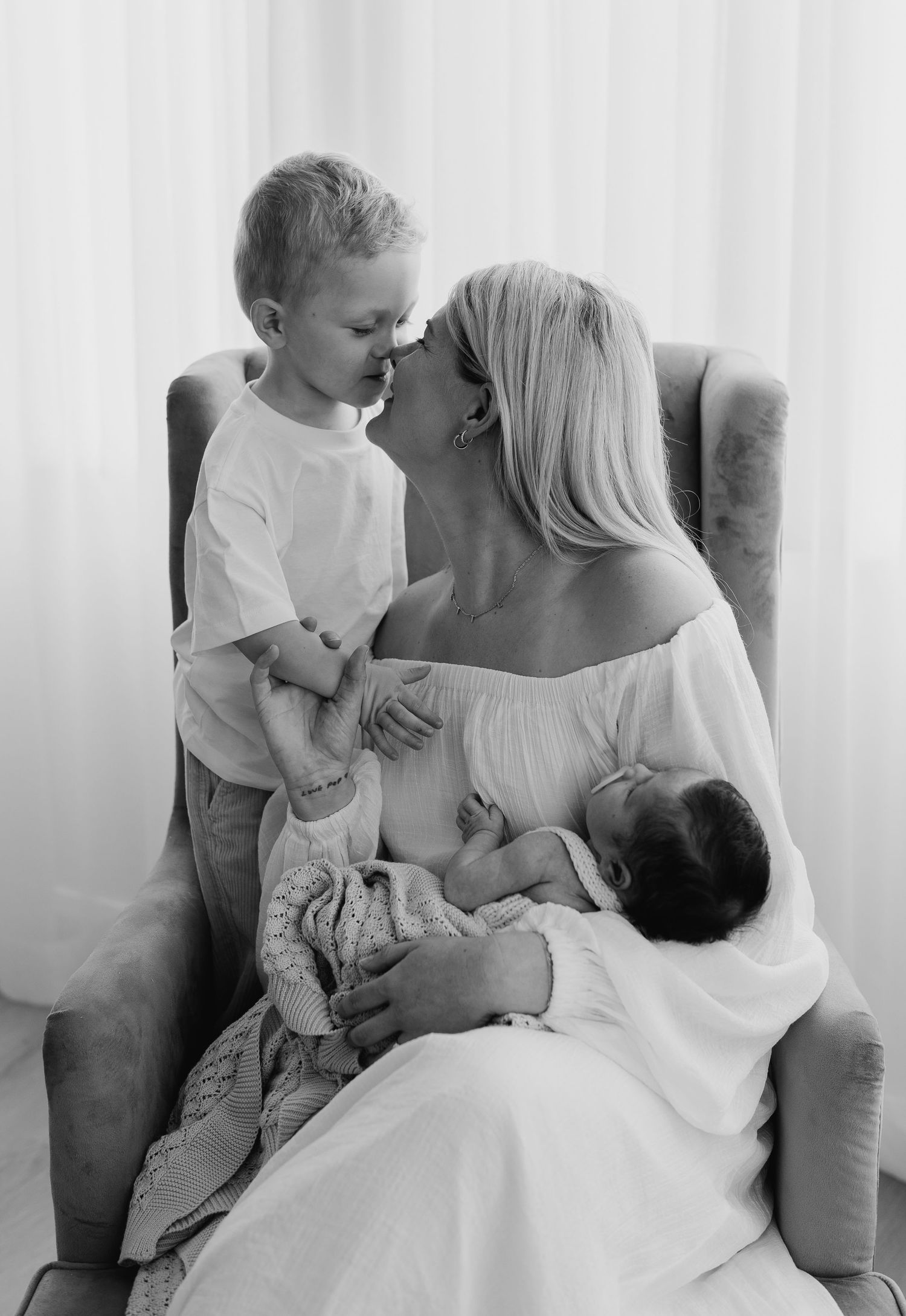 Black and white photo of a mother kissing her toddler while holding her newborn in a chair.
