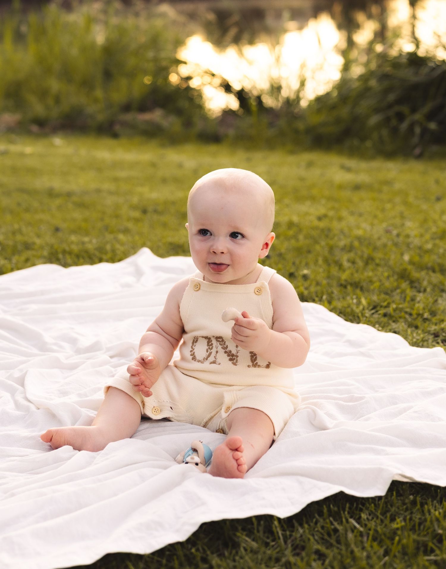 A baby in a cream romper sits on a white blanket on green grass, smiling outdoors in warm golden light.