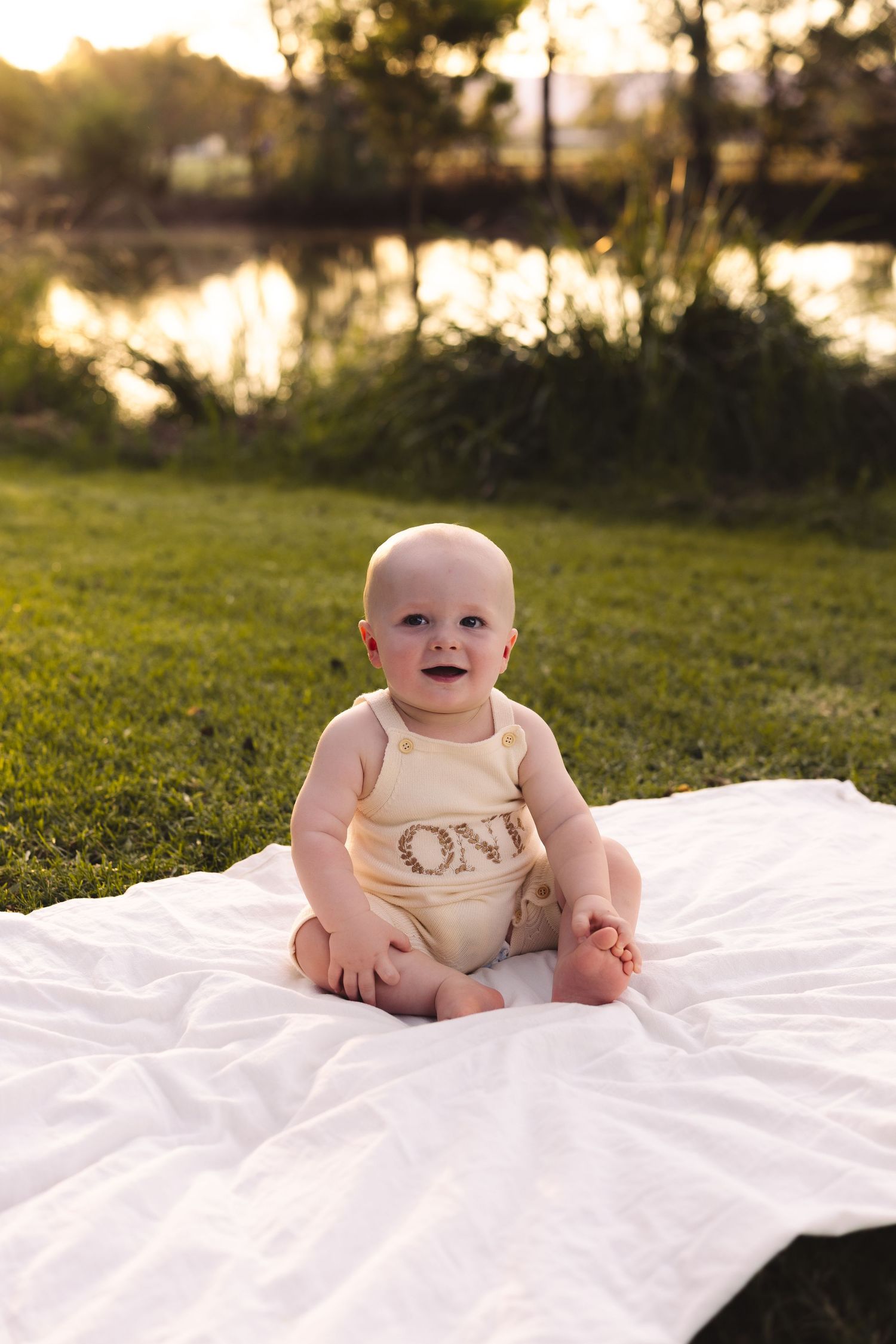 A smiling baby wearing a beige ONE romper sits on a white blanket in a sunny green park near a pond.