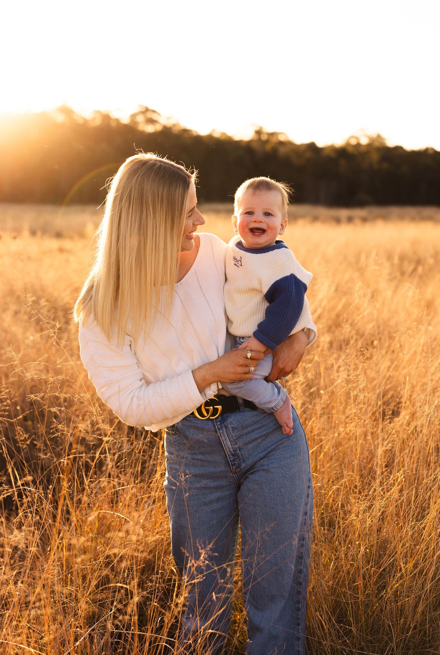 Blonde mother in white top holding laughing baby in golden grass field during warm sunset.