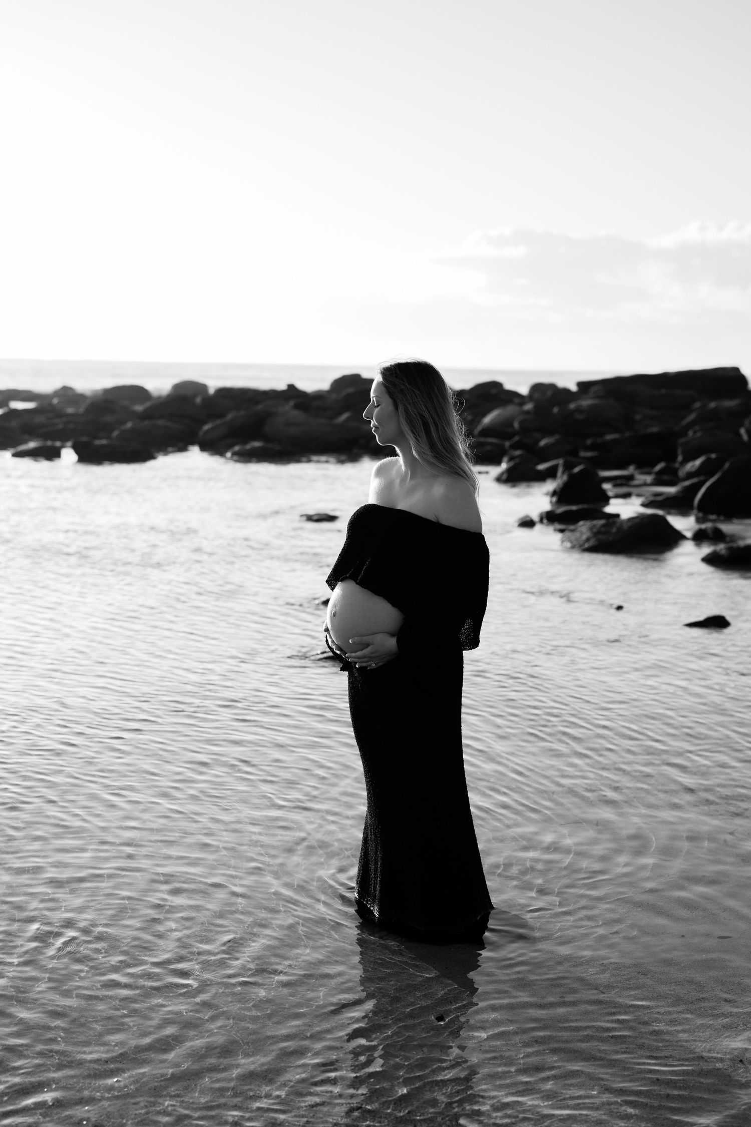 Black and white maternity photo of pregnant woman in black dress standing in shallow ocean water.