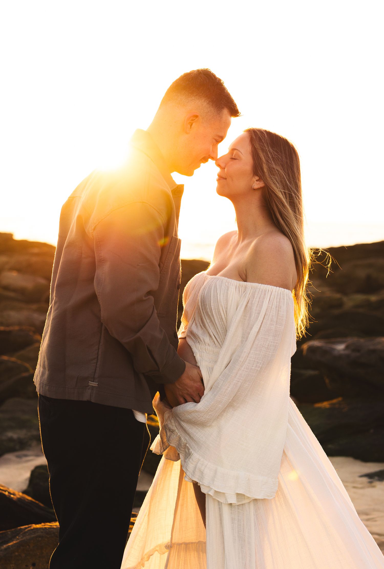 Couple touching noses romantically at sunset on rocky beach, woman in white off-shoulder dress.