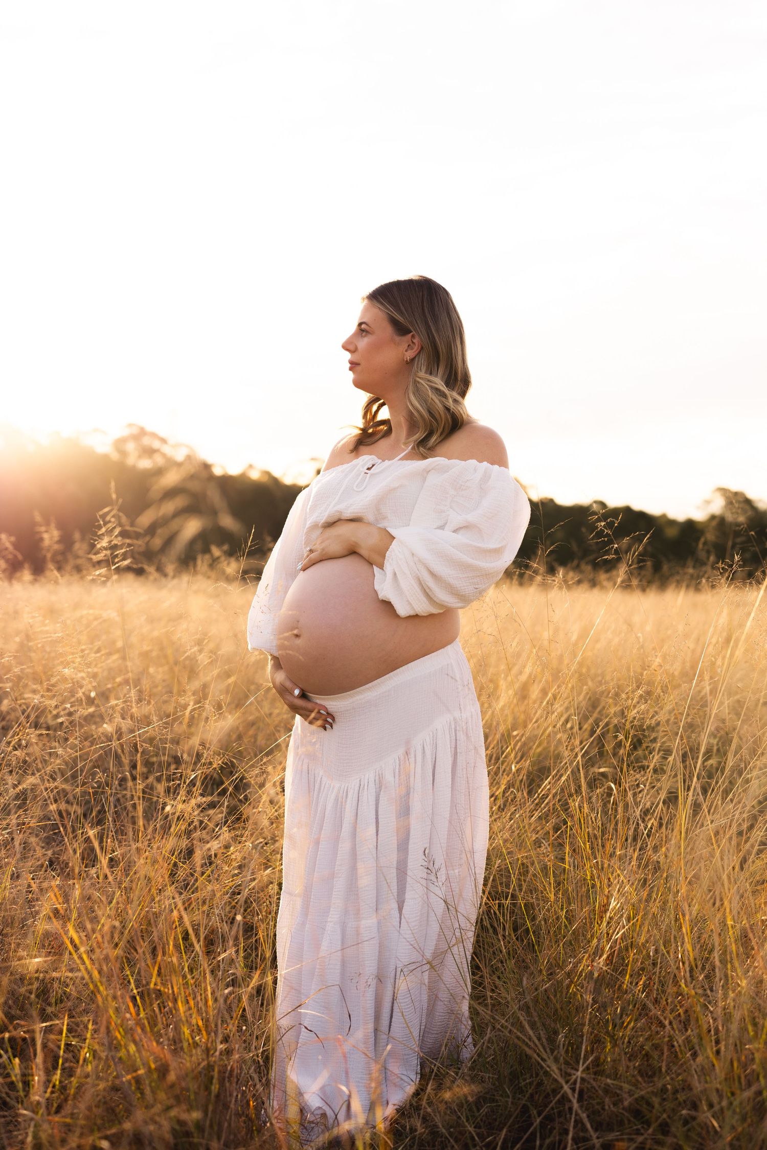 Pregnant woman in white off-shoulder dress cradling baby bump in golden field at sunset.