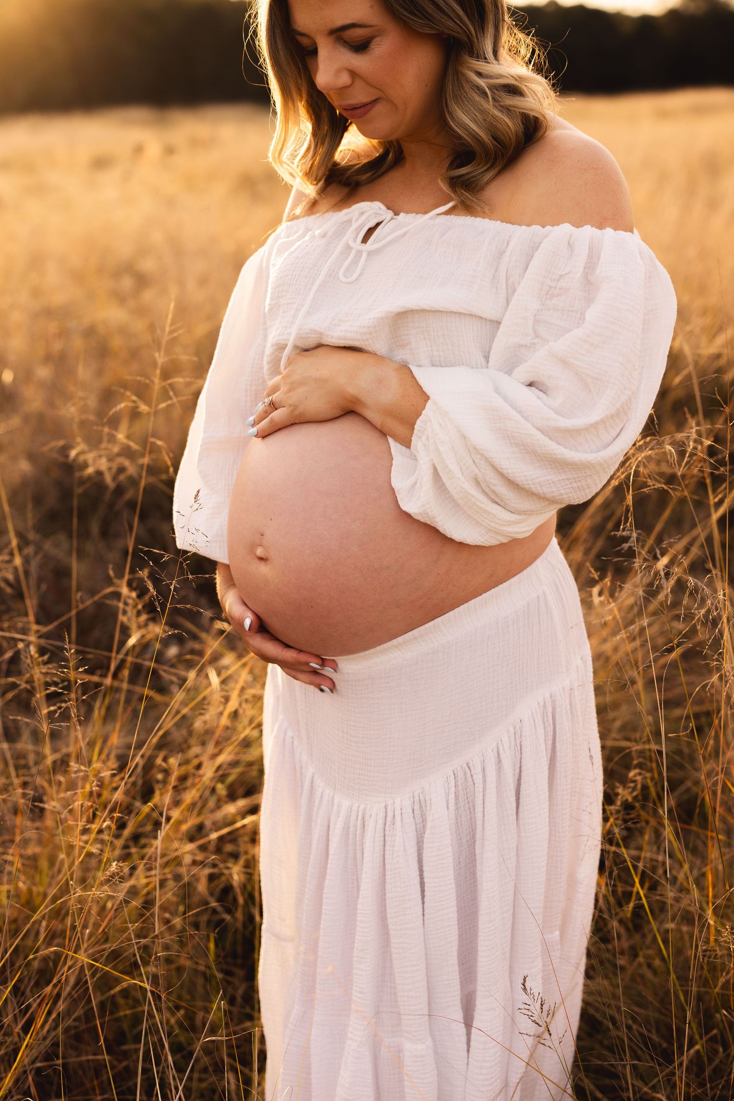 Pregnant woman in white off-shoulder dress lovingly cradling her bare bump in golden sunset field.
