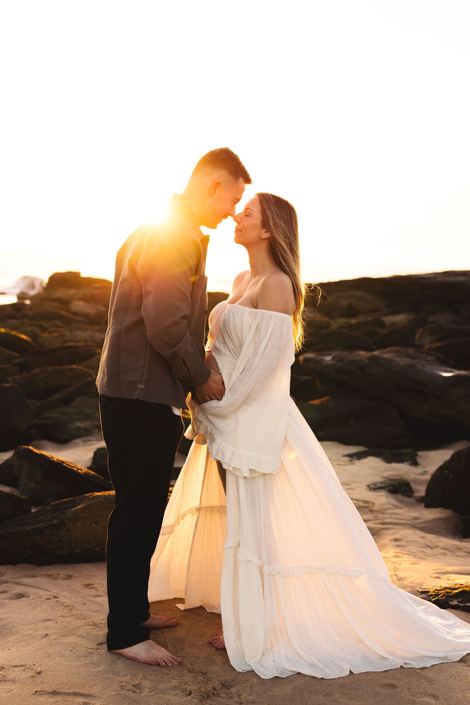 Couple sharing romantic moment at sunset on rocky beach, woman in flowing white dress.