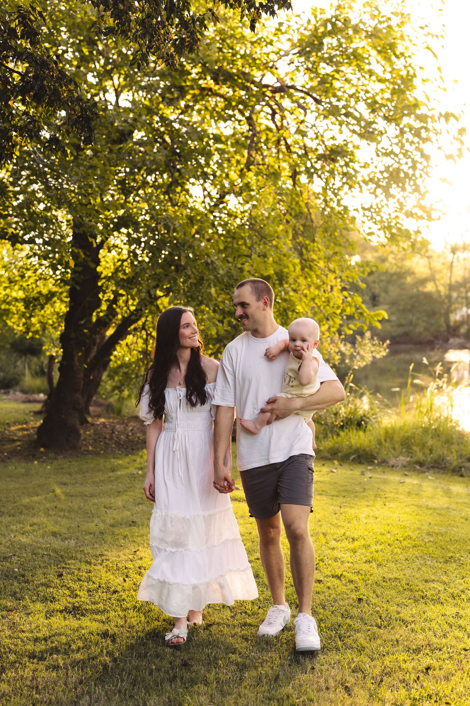 A happy young family with a baby enjoys a golden hour stroll on lush green grass beneath a sunlit tree.