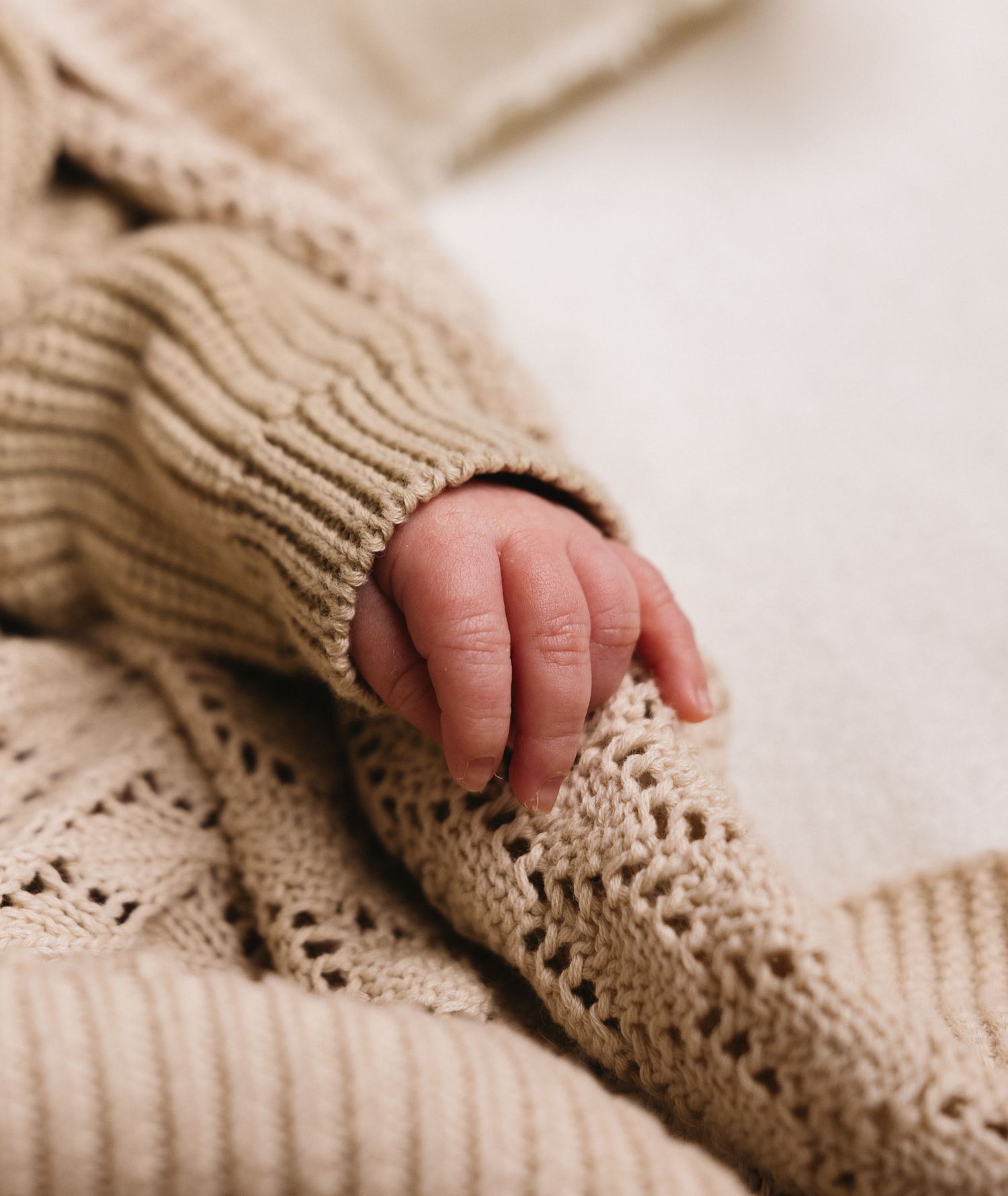 Close-up of newborn baby tiny hand peeking out from a soft beige knitted sweater on cream blanket.