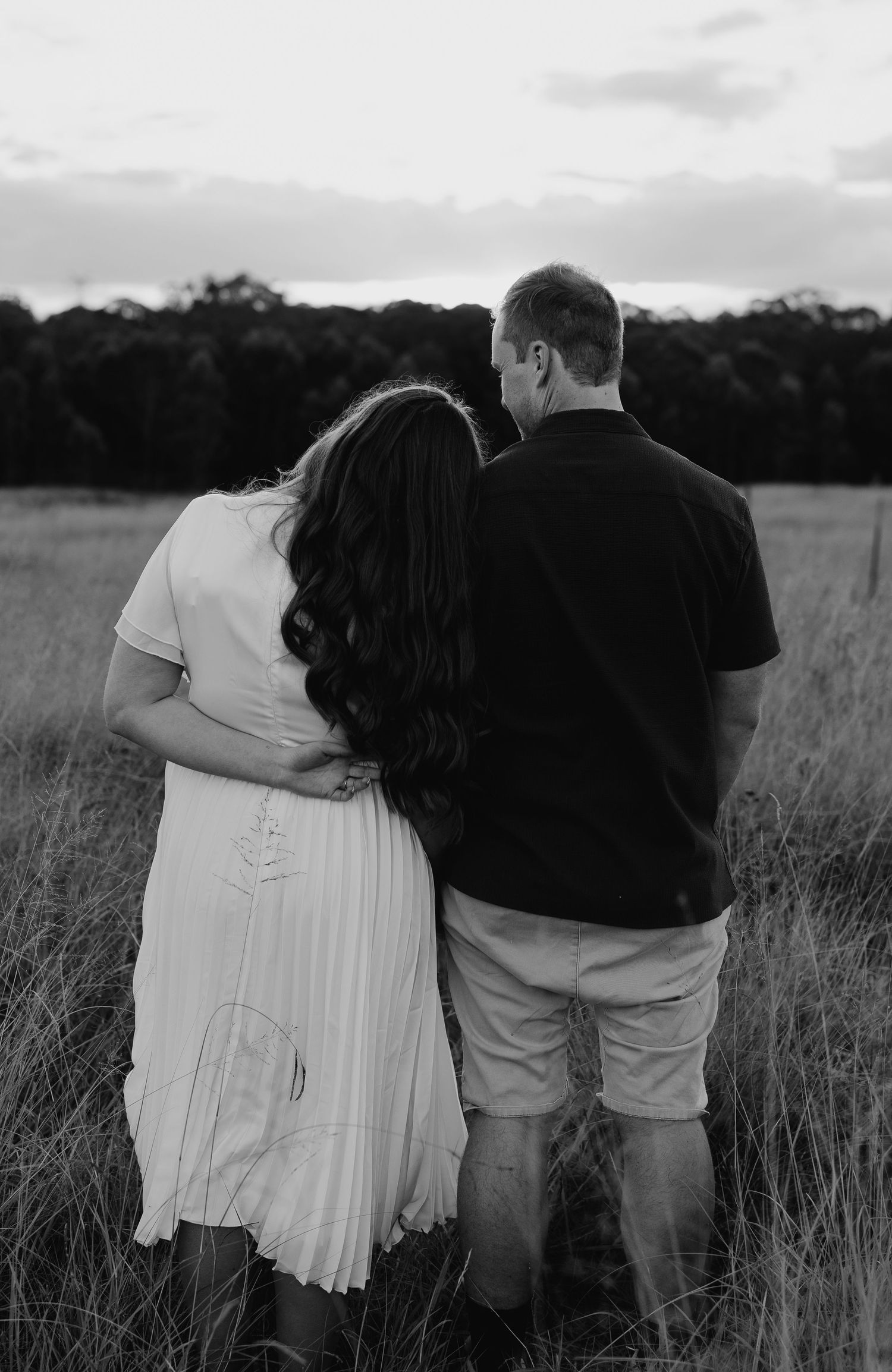 Black and white photo of couple standing arm in arm facing away in grassy field at sunset, woman in white dress.