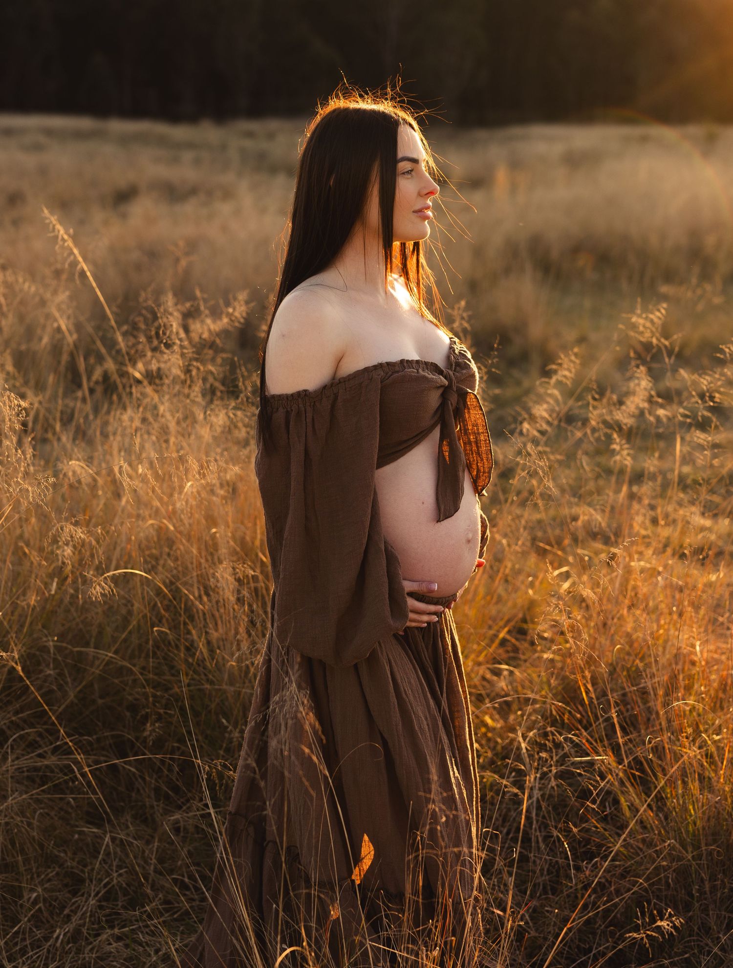 Pregnant woman in brown off-shoulder dress holding belly in golden field at sunset.