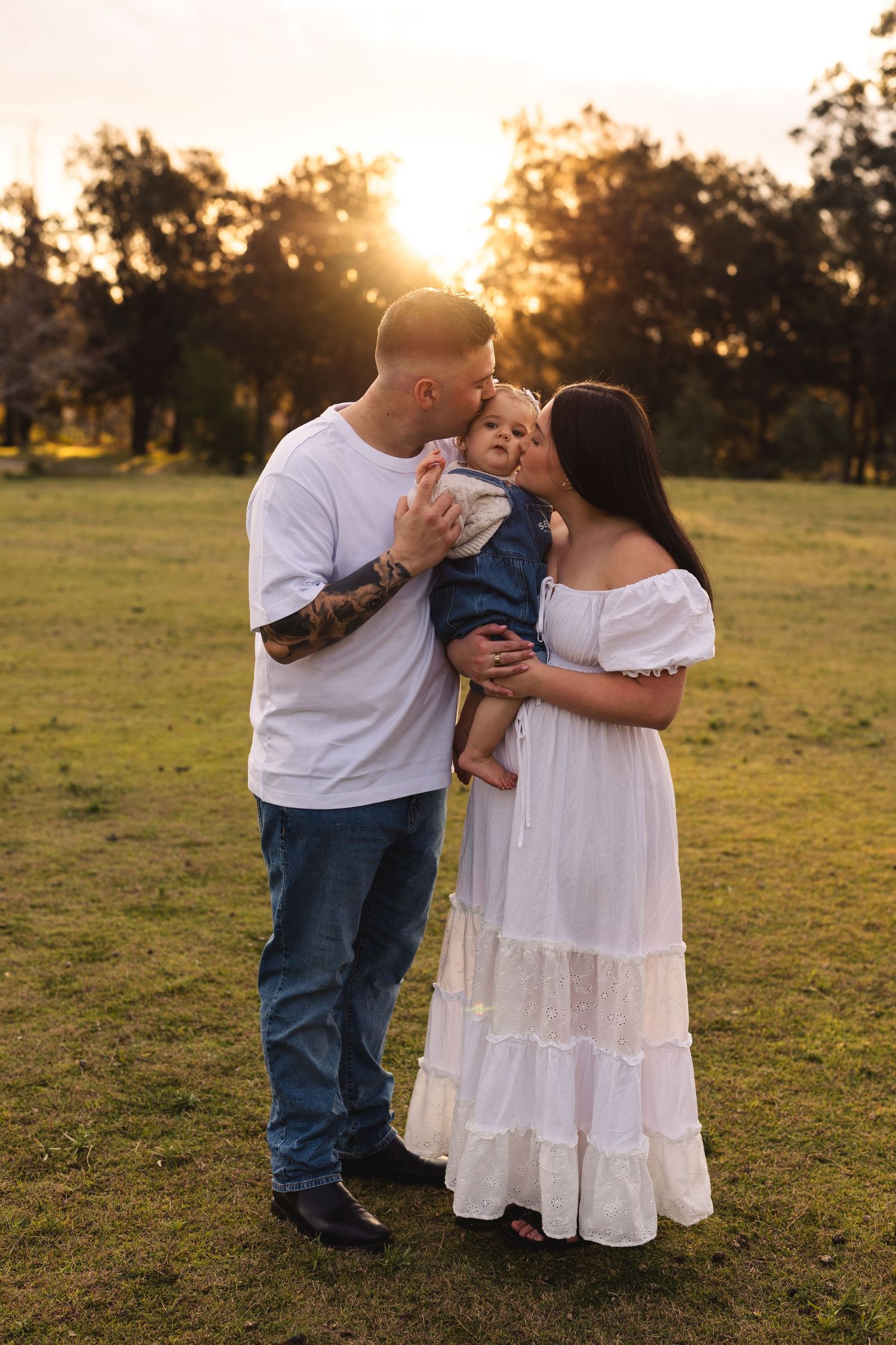 A young family shares a tender moment at sunset in a park, with dad kissing baby as mom smiles in a white dress.
