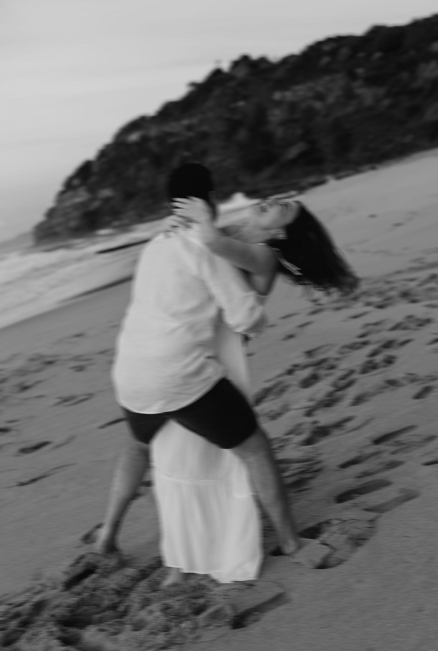 Black and white blurred artistic photo of couple sharing dramatic dip kiss on sandy beach at dusk.