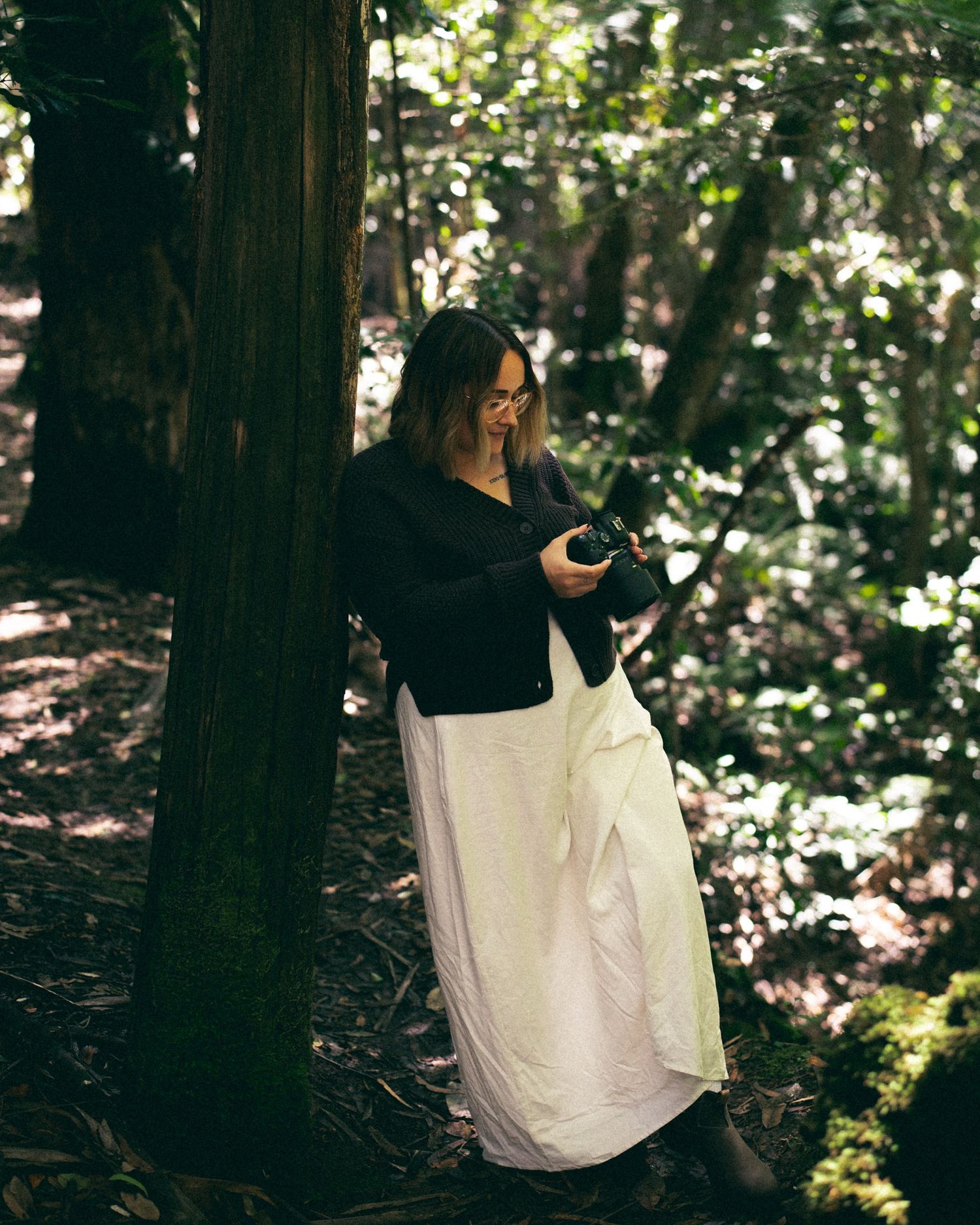 Woman in black top and white skirt reviewing photos on camera in forest.