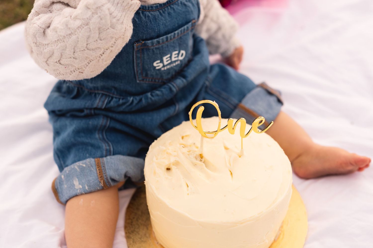 Baby in denim overalls sits beside a first birthday smash cake with gold 