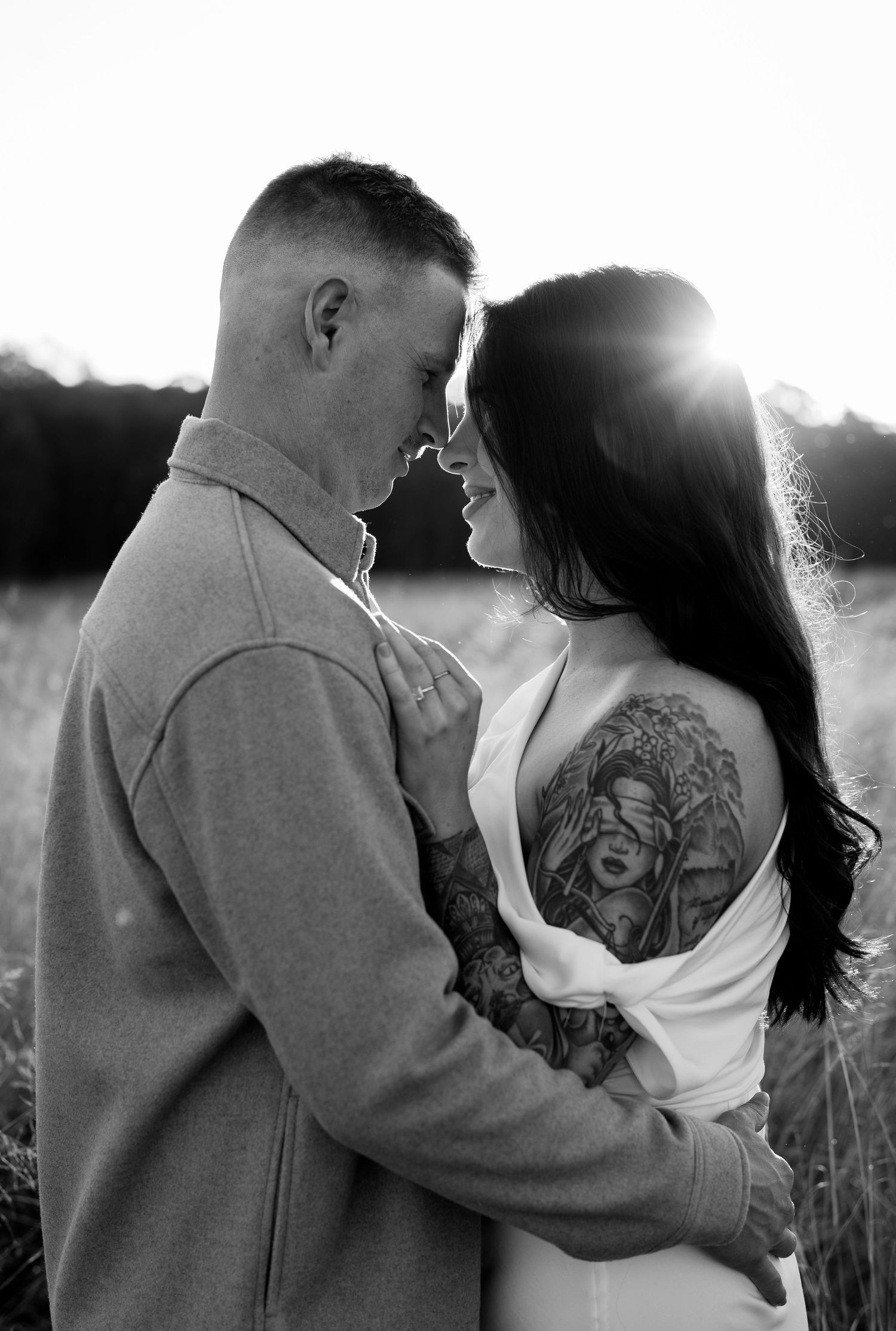 Black and white intimate couple embracing at sunset, woman with large back tattoo in white top, man in grey jacket.