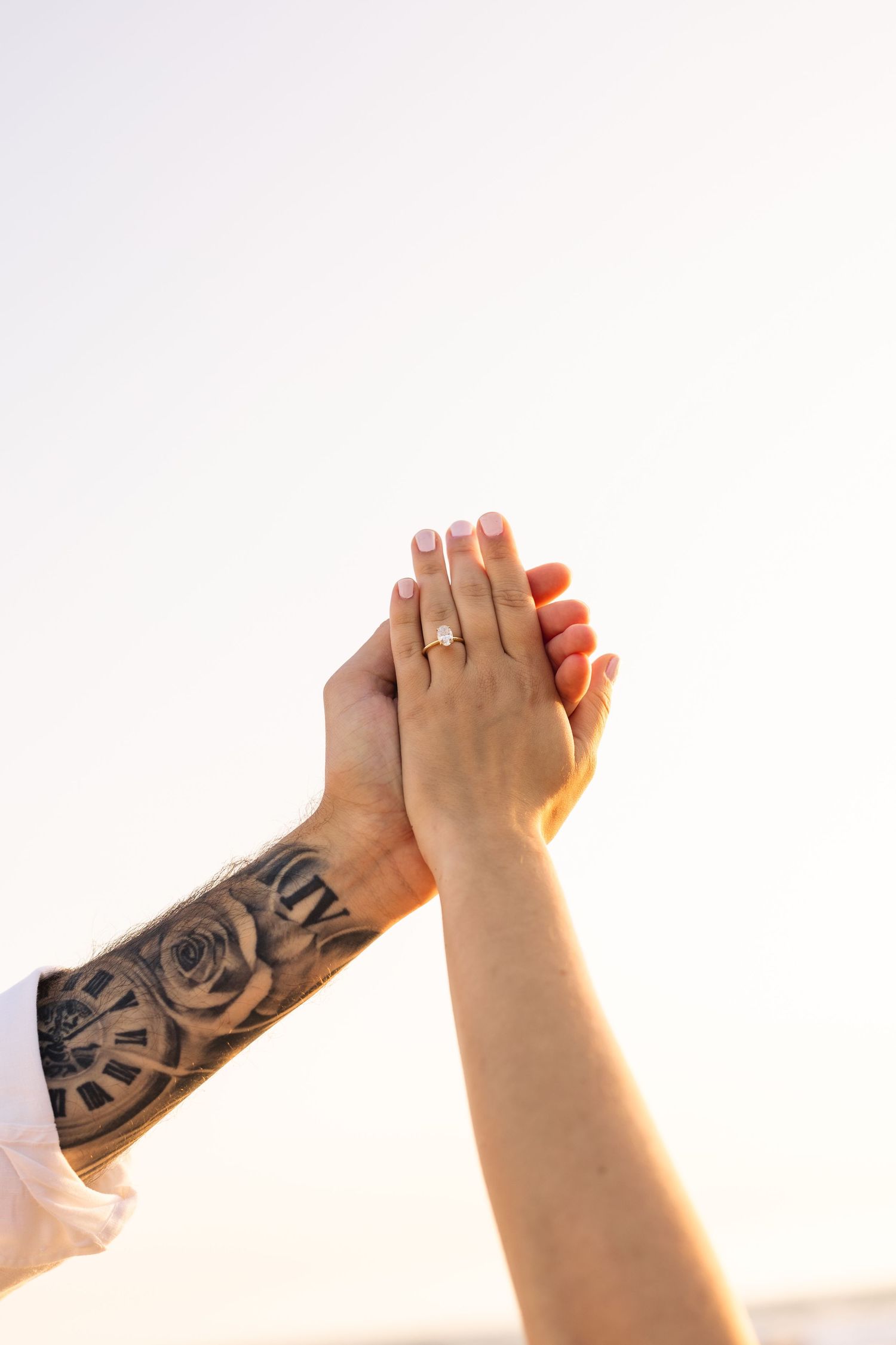 Two hands raised against sky, one tattooed, the other wearing a diamond engagement ring.