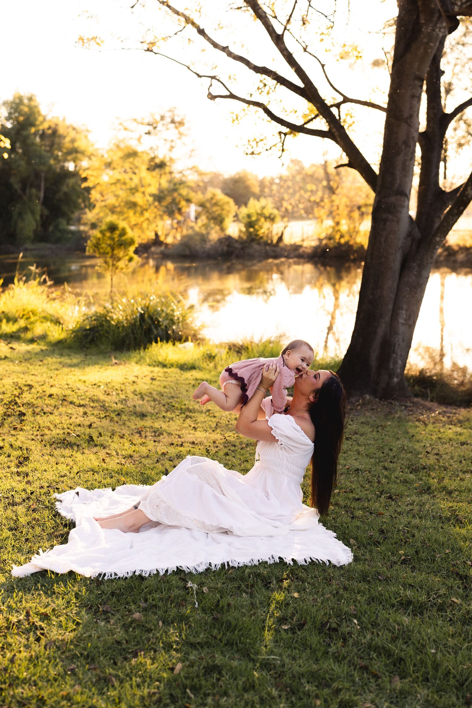 Mother lying on blanket lifting smiling baby by serene lakeside at sunset.