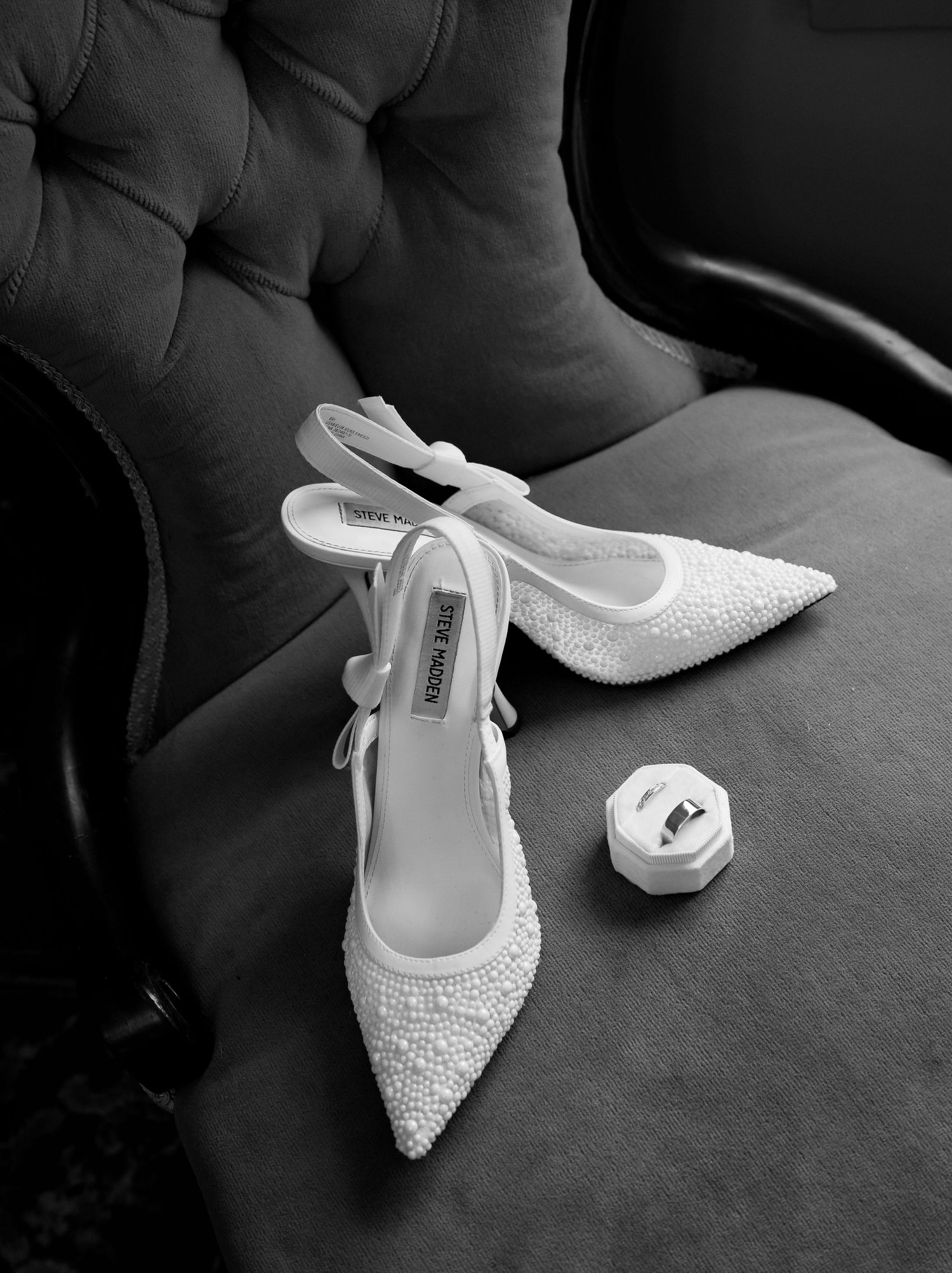 Black and white flat lay of pearl bridal heels and ring box on velvet chair.