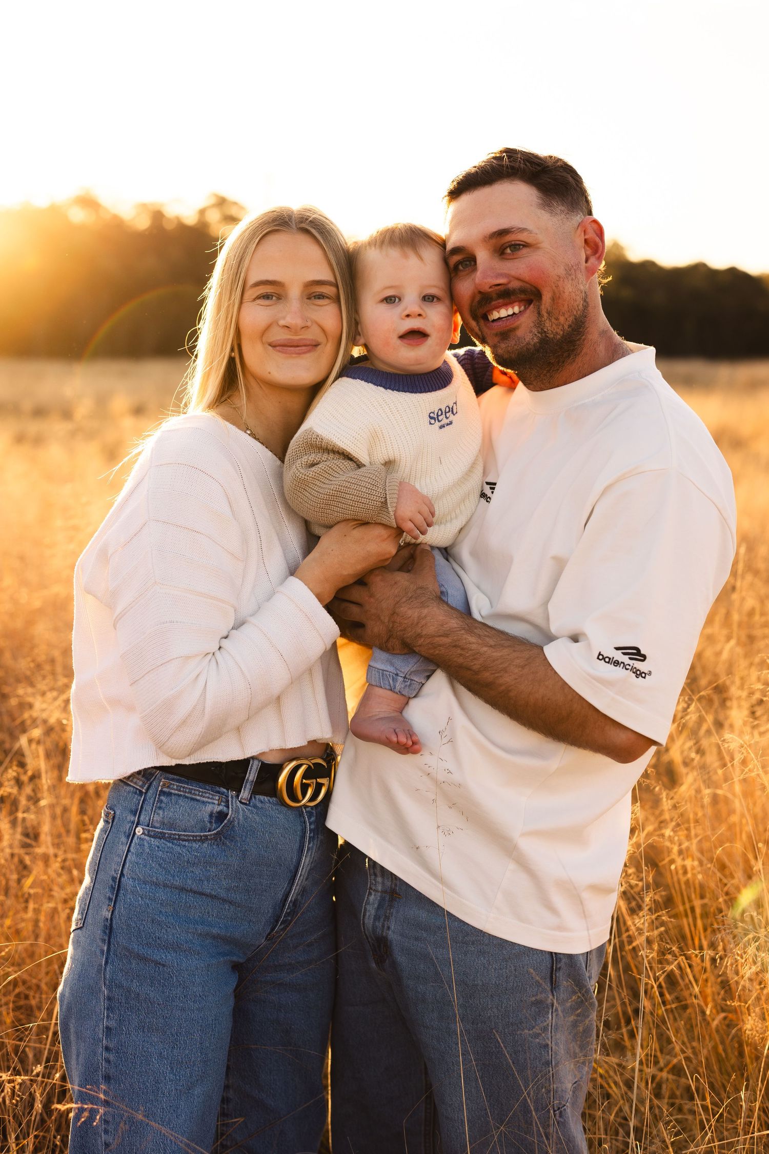 Young family with baby posing in golden wheat field at sunset, warm glowing light behind them.