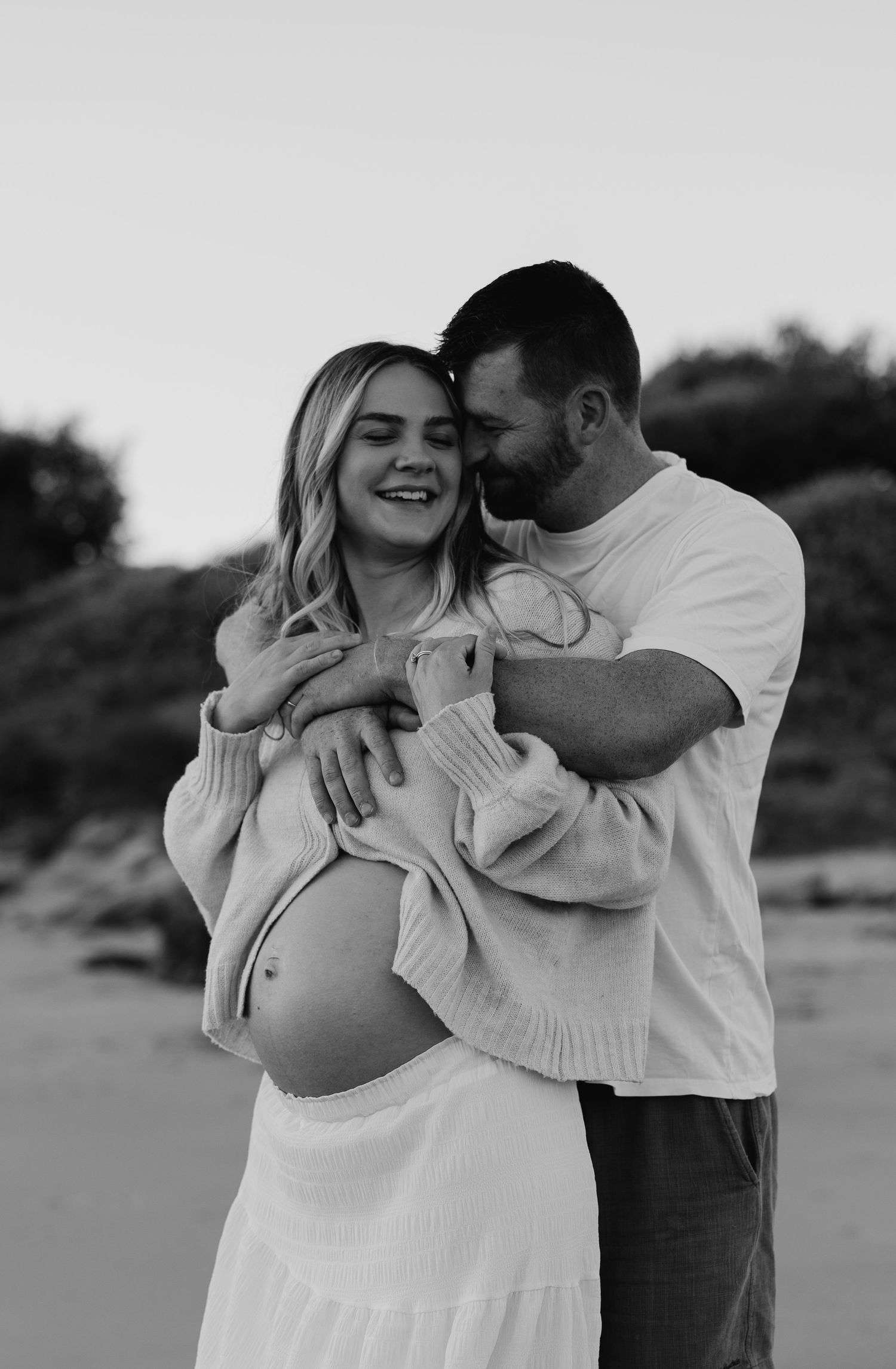 Black and white photo of smiling pregnant woman embraced by partner on beach, both cradling her baby bump tenderly.