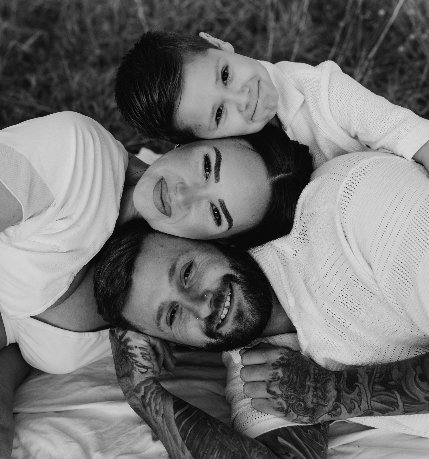 Black and white family portrait of smiling parents and child stacked together, man with tattoos, outdoors.
