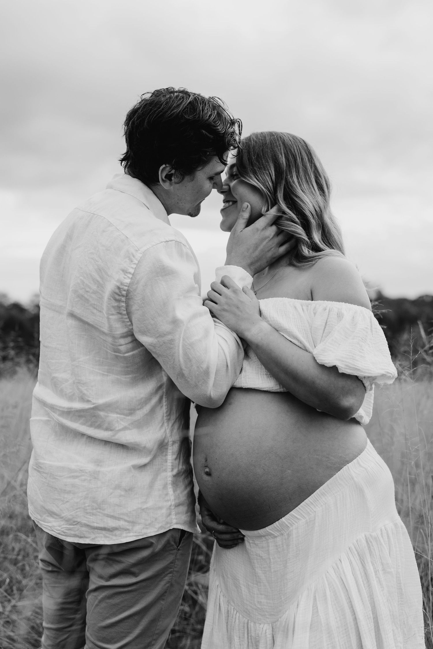 Black and white photo of couple touching foreheads tenderly outdoors, woman in white dress showing pregnant belly.
