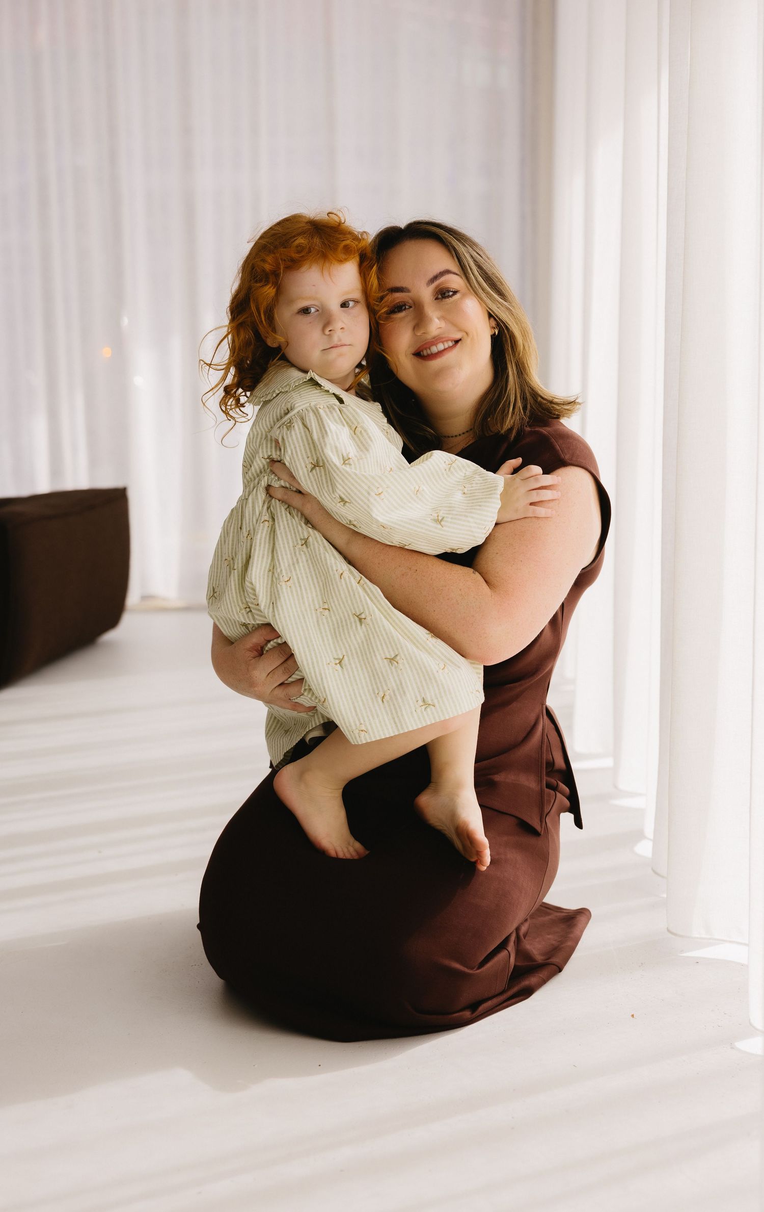 Smiling mother kneeling and hugging her red-haired toddler daughter in a bright white room.